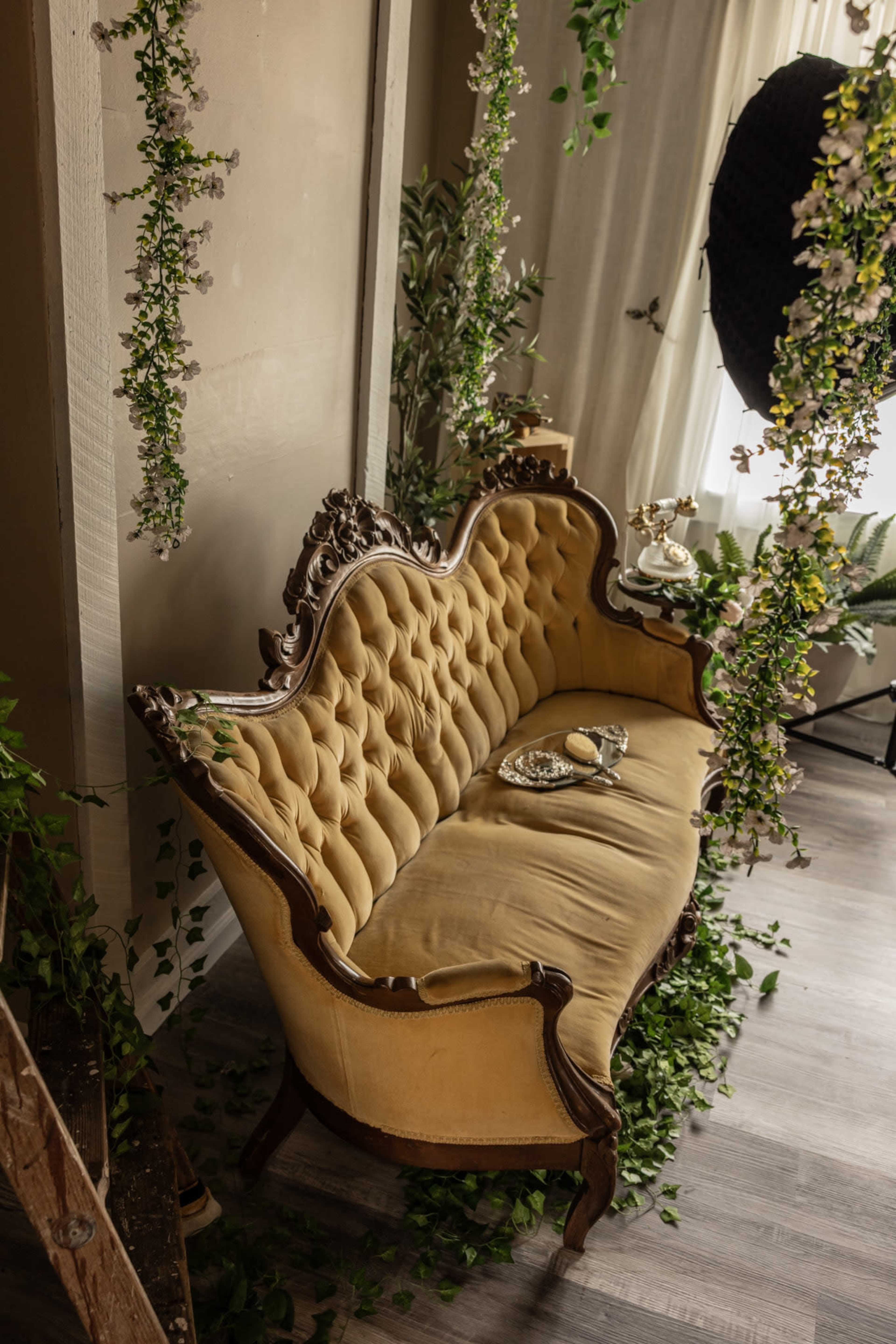 A vintage yellow tufted sofa with ornate woodwork is positioned in a room adorned with hanging greenery and floral accents.