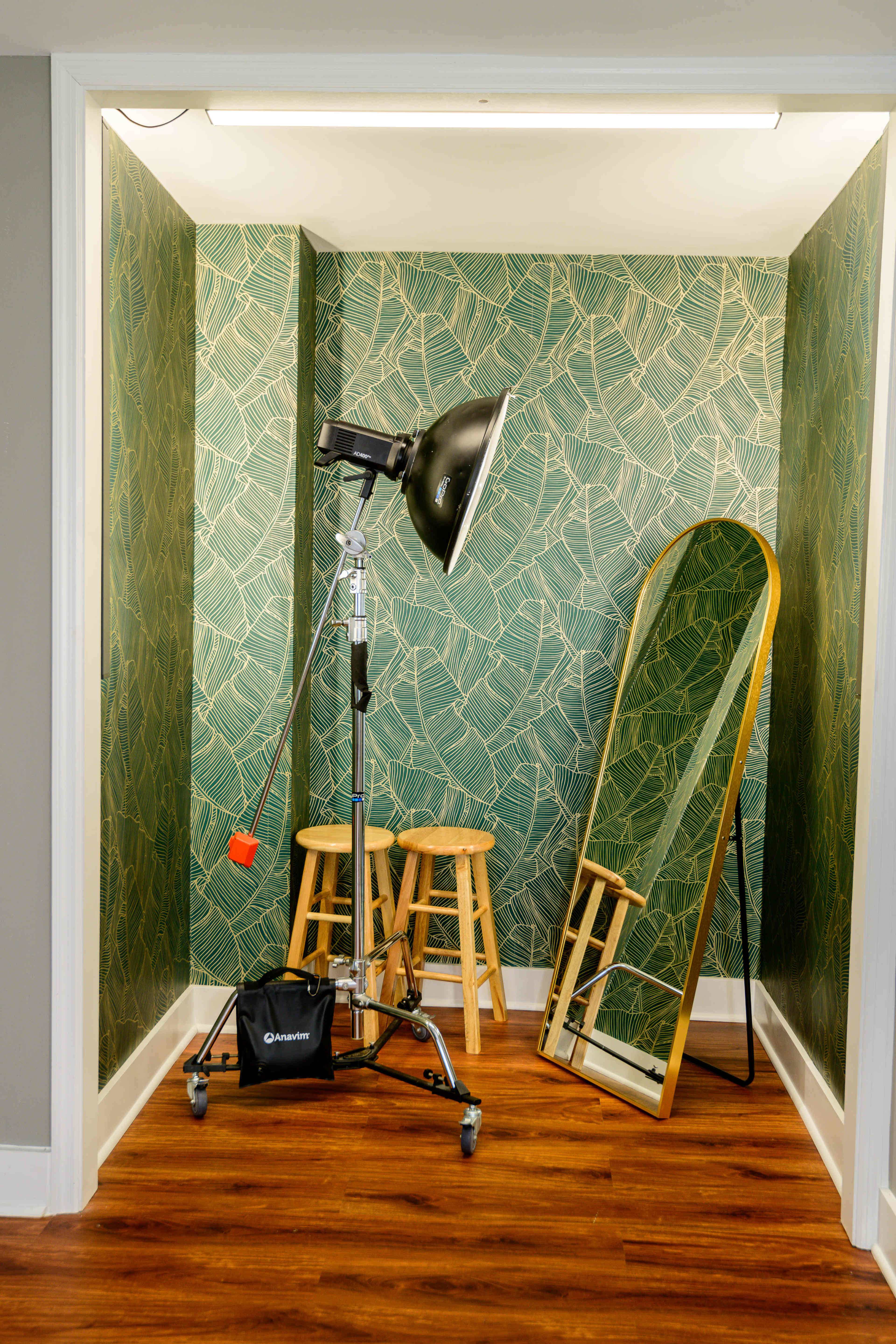 A small room features a green patterned wallpaper, a full-length mirror, three wooden stools, and a floor lamp on a stand.