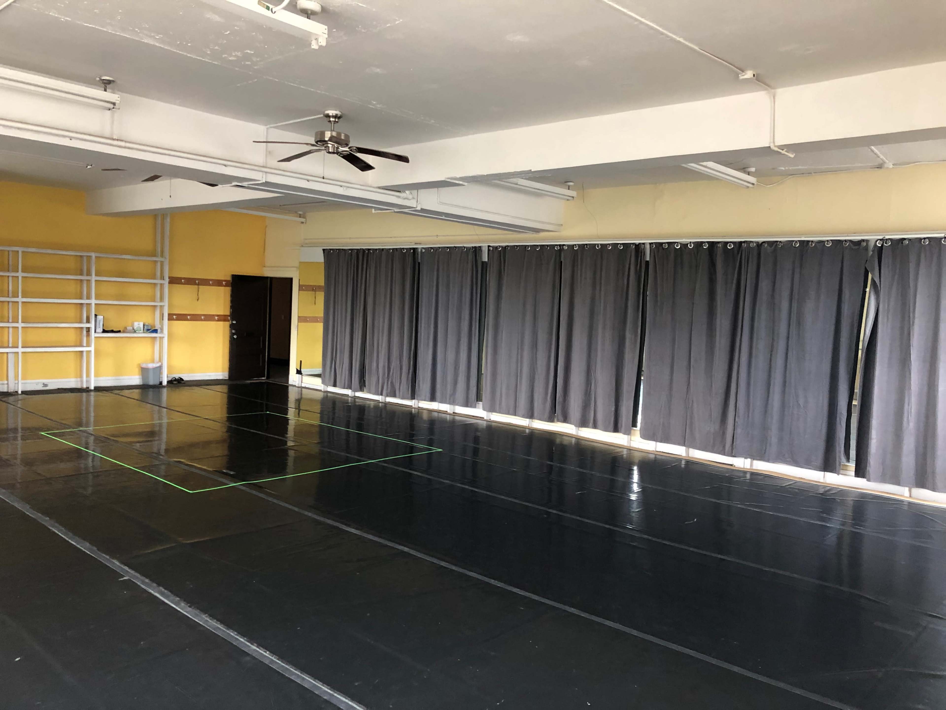 A spacious dance studio with black flooring, gray curtains covering the windows, and yellow walls.