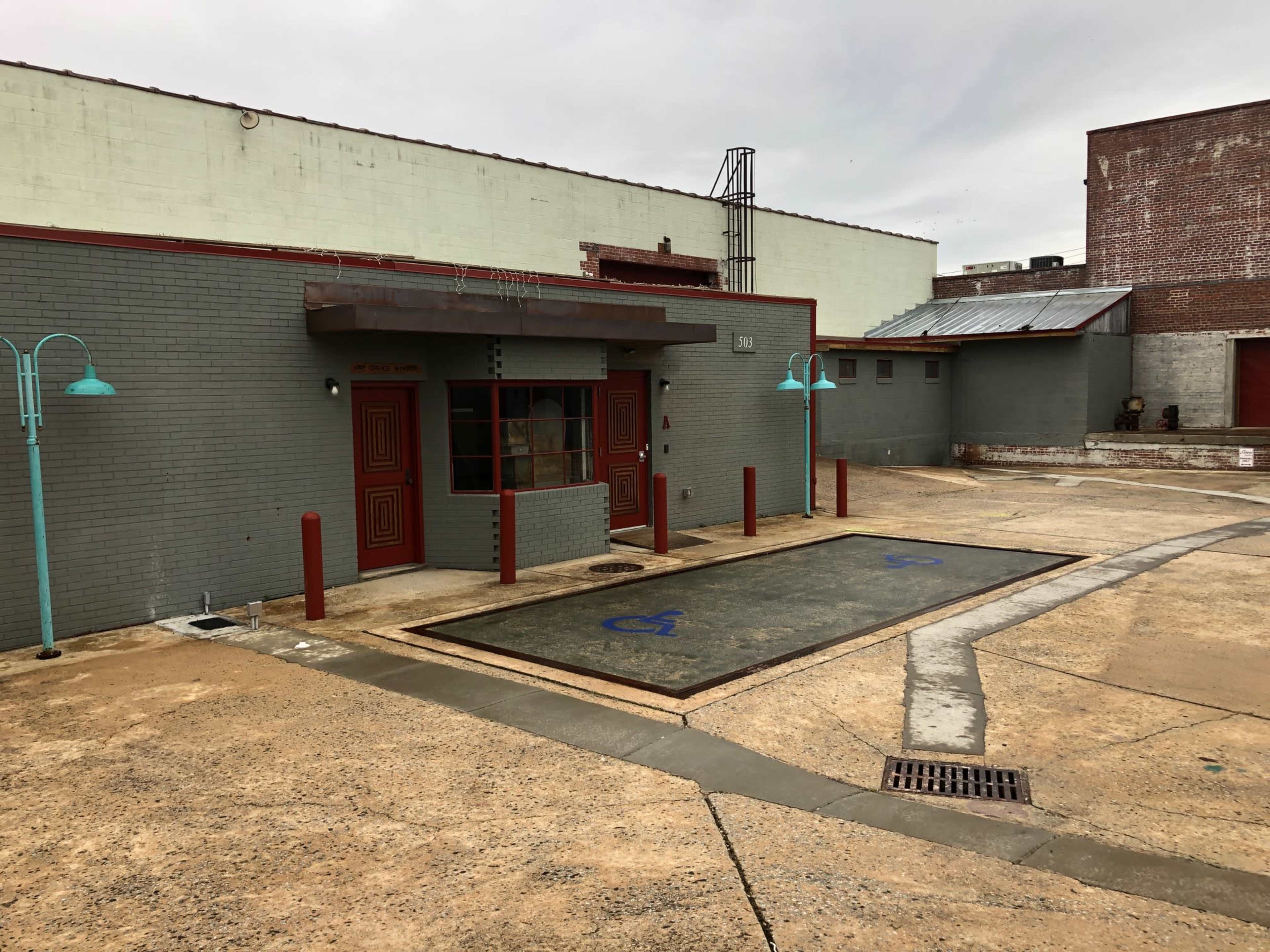 The image shows a gray, one-story building with a front entrance, a blue handicap-accessible parking space, and light blue lamp posts in a concrete area.