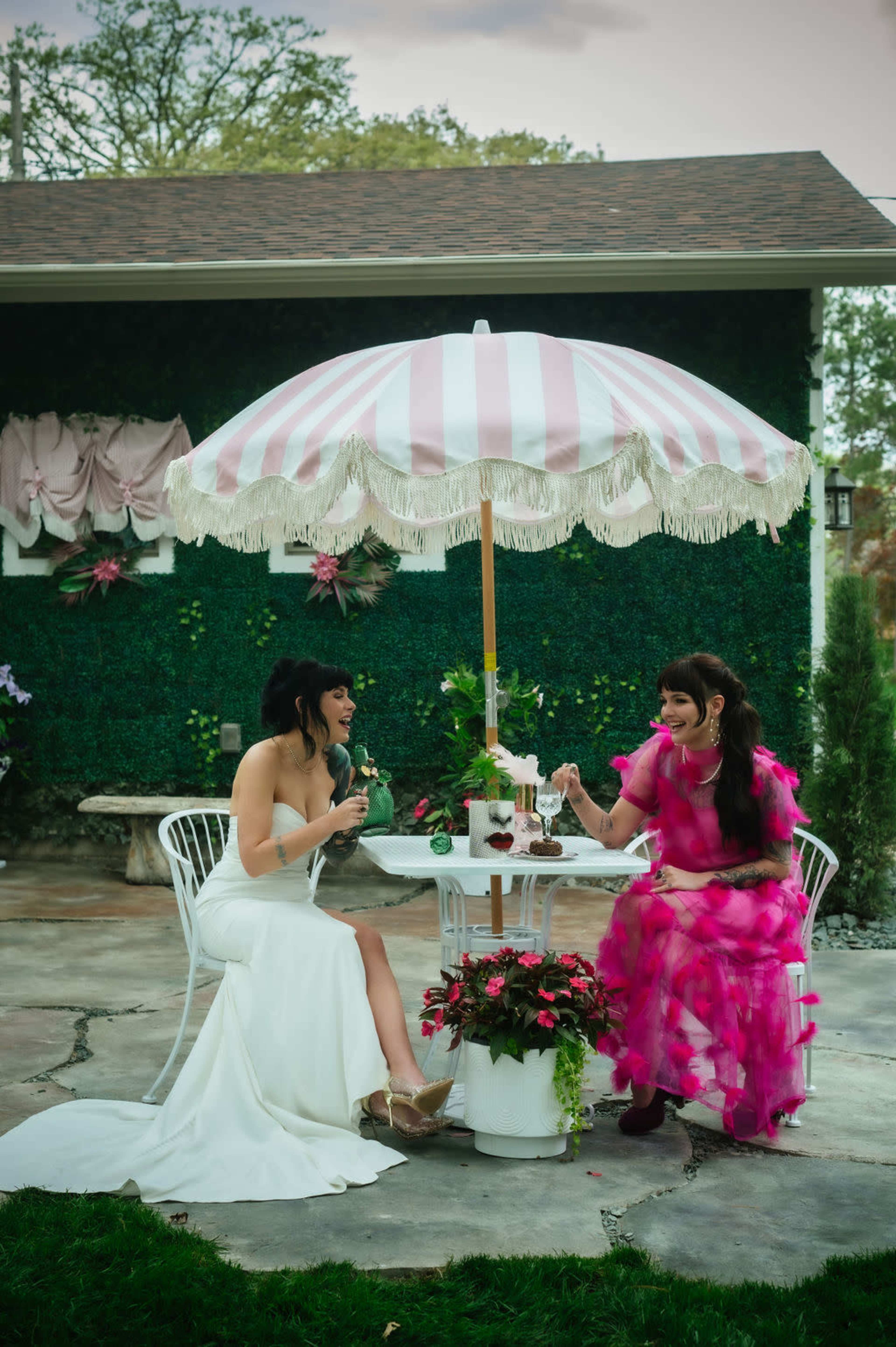 Two women sit under a pink and white striped umbrella at a table adorned with flowers, enjoying refreshments in a garden setting.