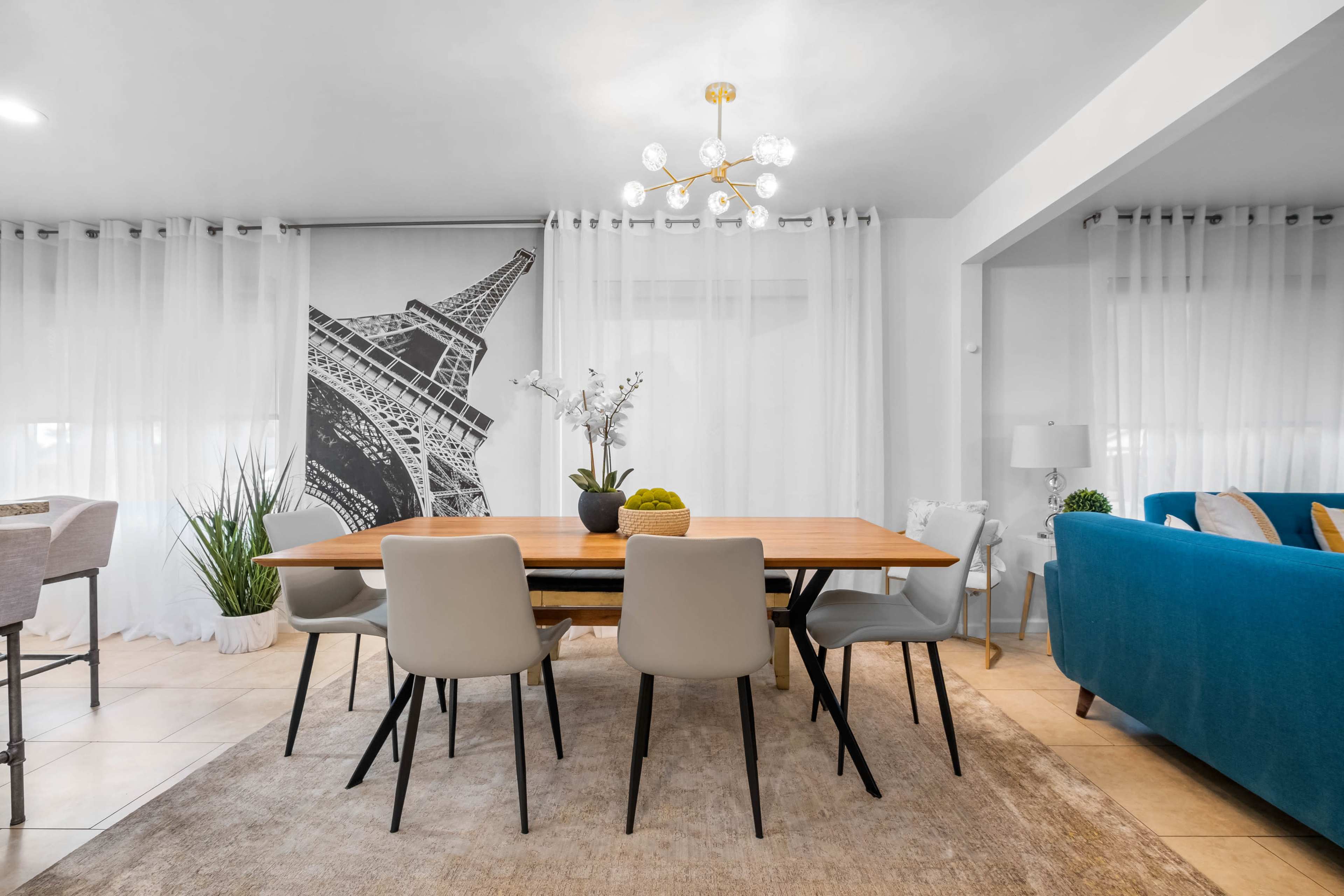 A dining area features a wooden table surrounded by chairs, with a backdrop of sheer curtains and a mural of the Eiffel Tower.