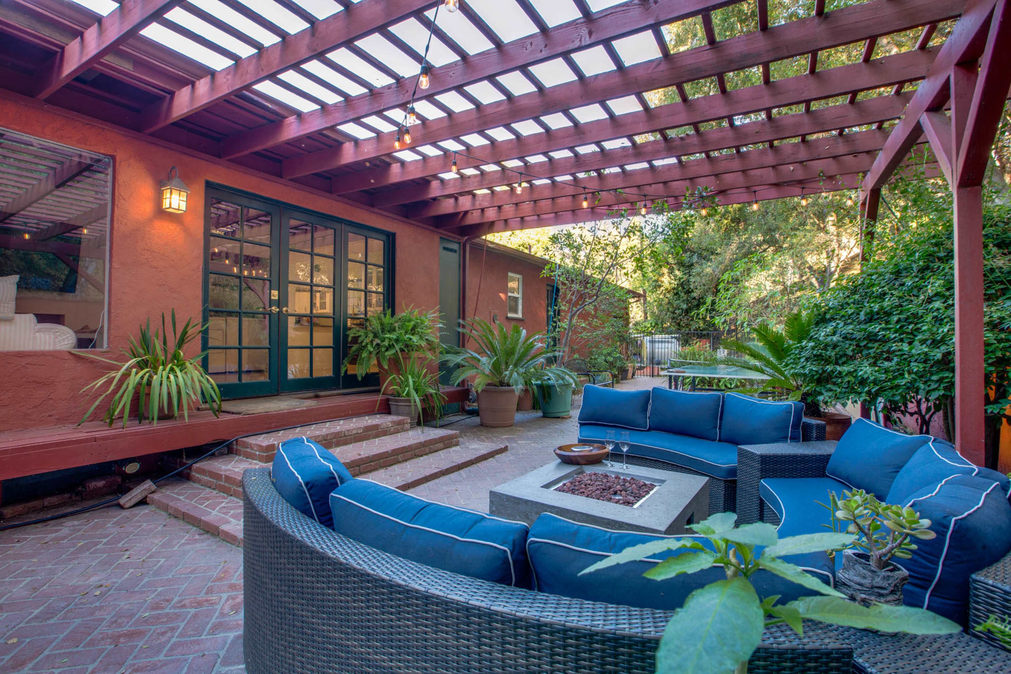 A cozy outdoor living space features a circular seating arrangement around a fire pit, surrounded by lush greenery and a wooden pergola.