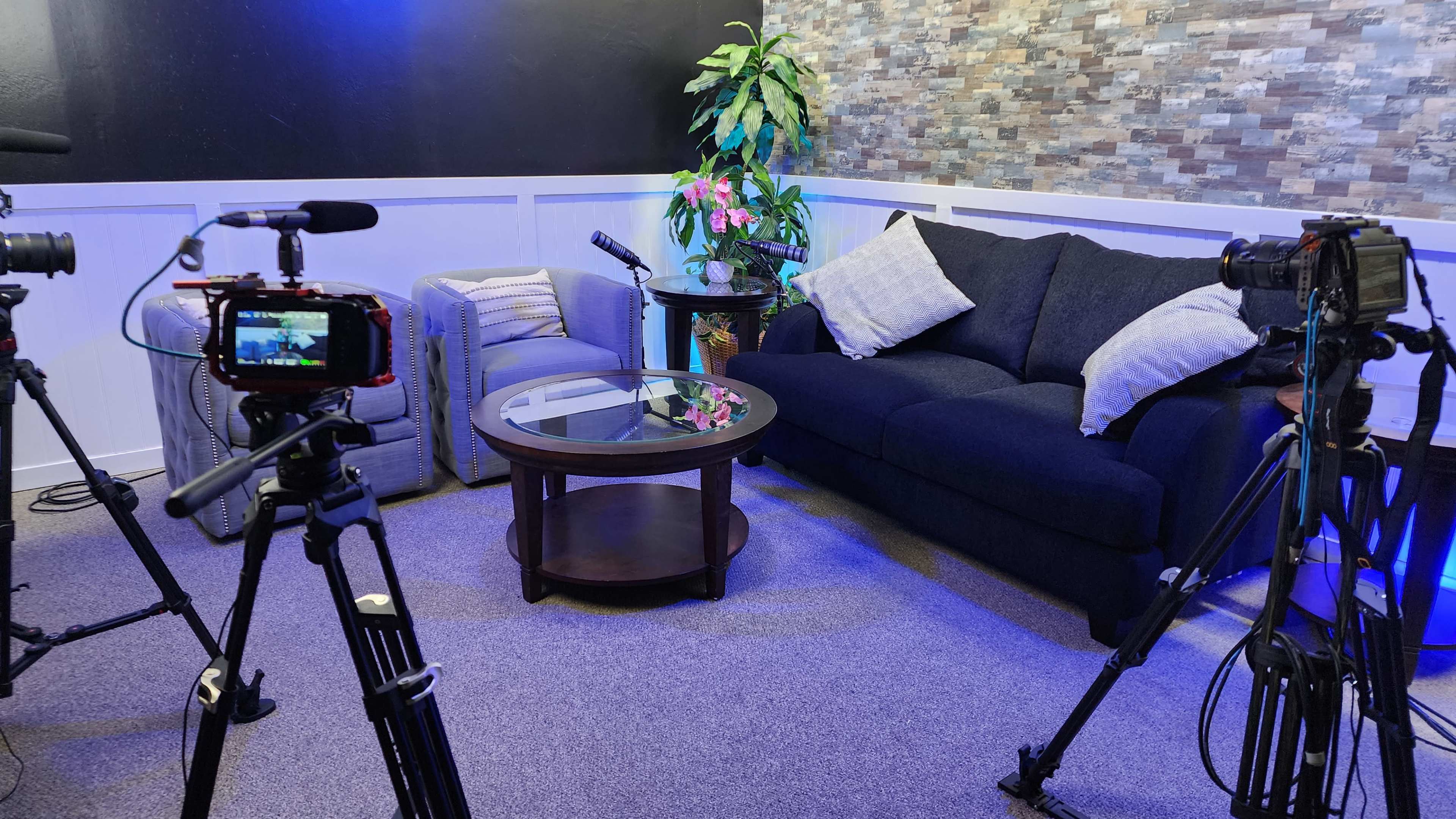 The scene features a cozy studio setup with a dark sofa and armchair, a coffee table in the center, and camera equipment positioned around the area.