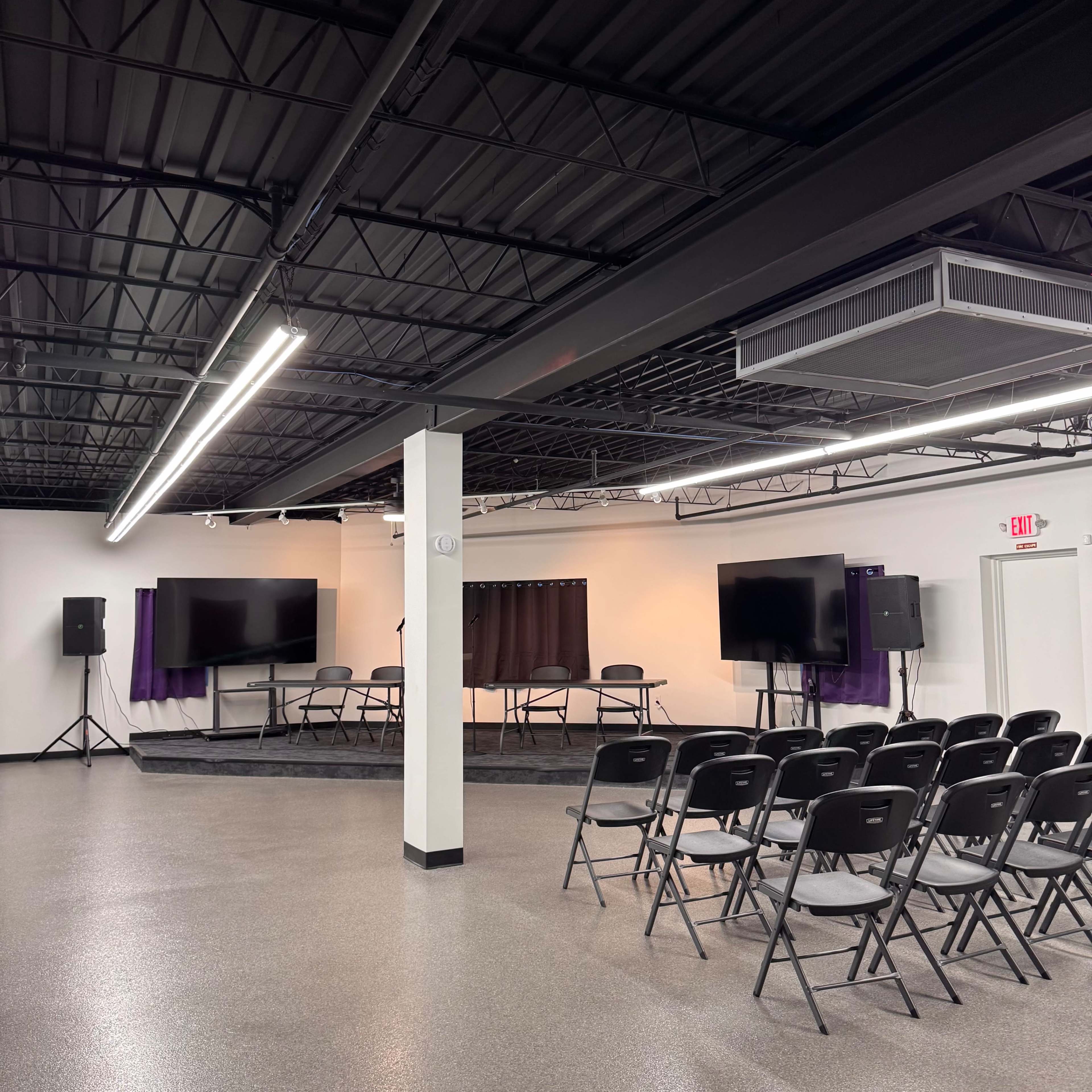 The image shows a spacious room set up for an event, featuring rows of black chairs facing a stage with two large screens and a panel table.
