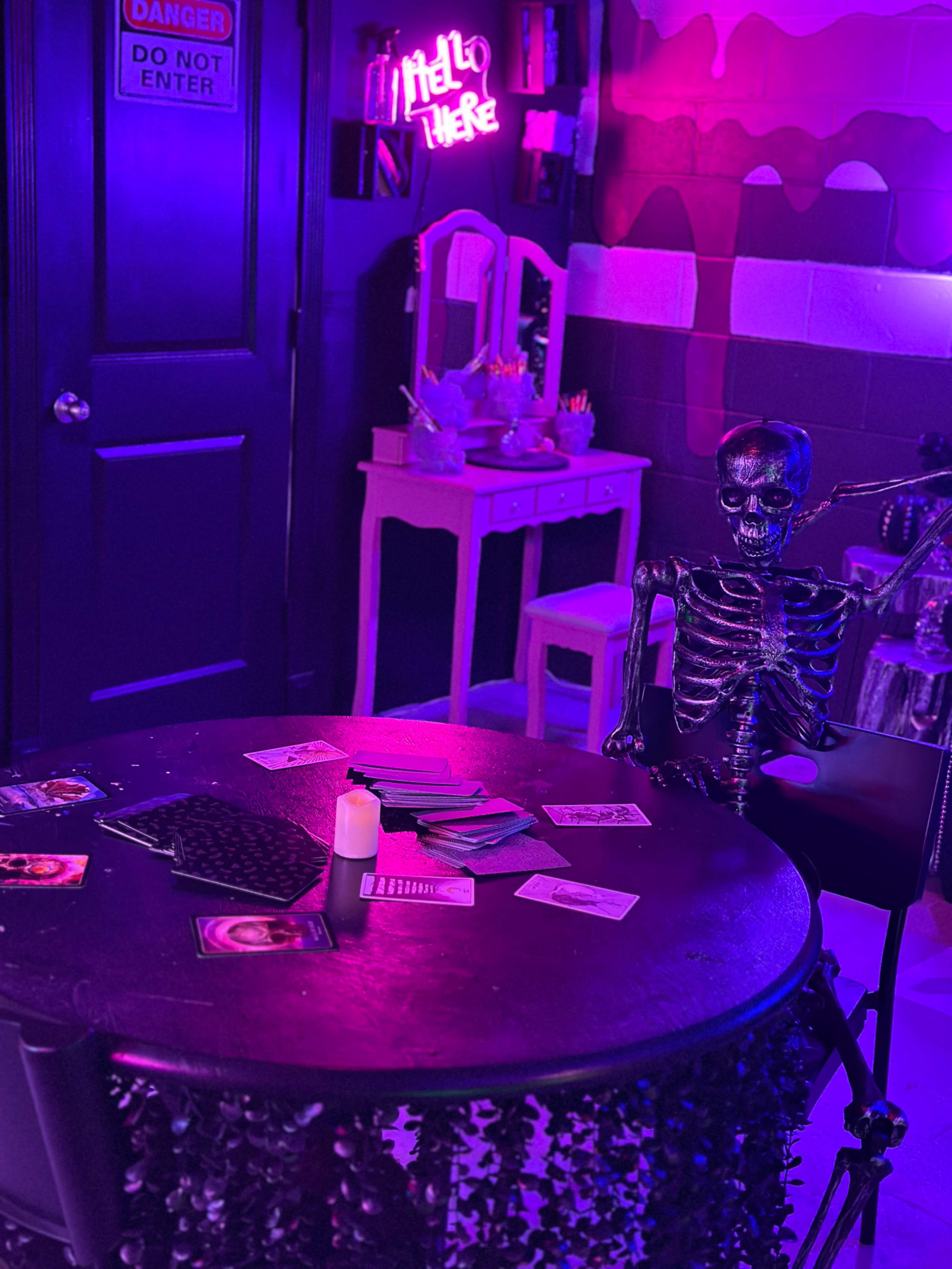 A dimly lit room features a table with playing cards, a candle, and a skeleton seated beside it, with purple neon lighting illuminating the space.