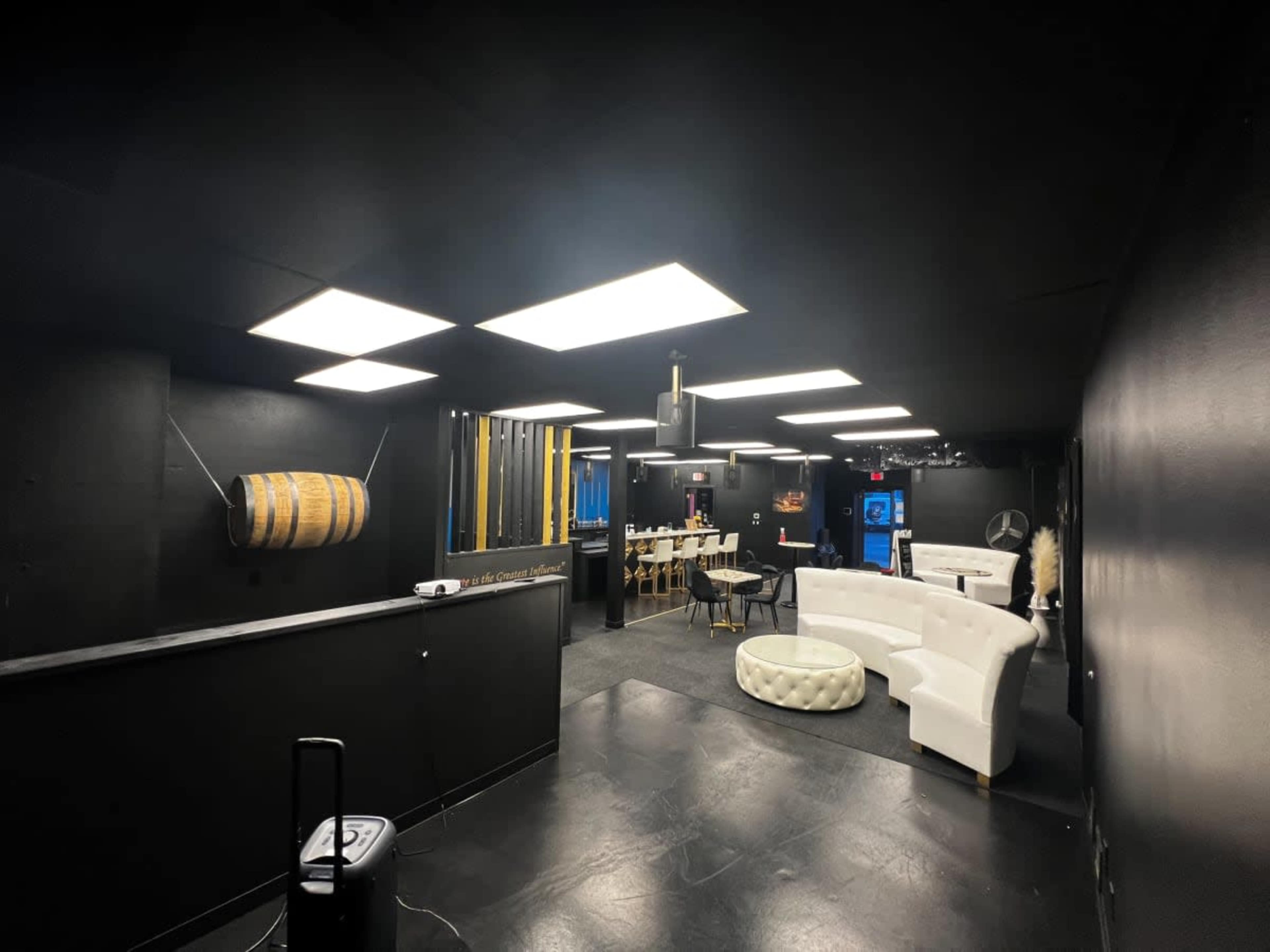 A dimly-lit lounge area with black walls, a white couch, a barrel on display, and a dining table set for several people.