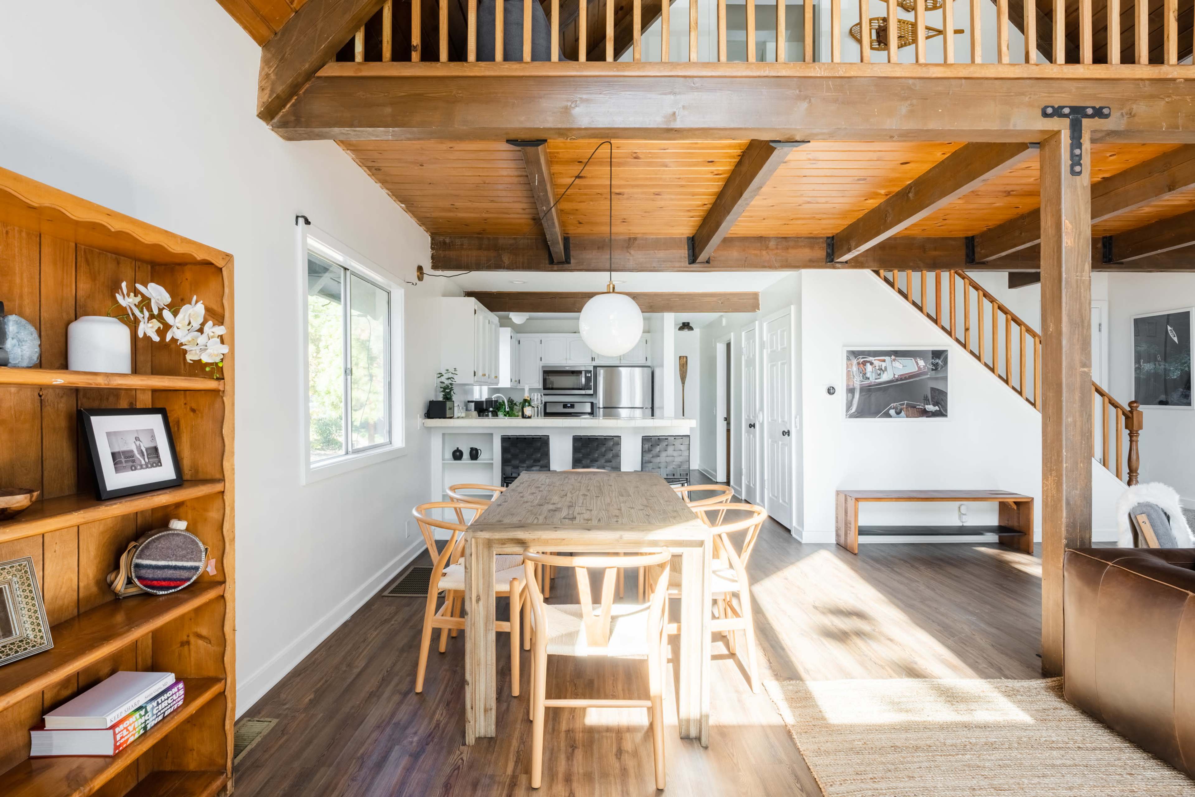 A spacious dining area features a wooden table surrounded by chairs, with a staircase leading to an upper level and an open kitchen in the background.
