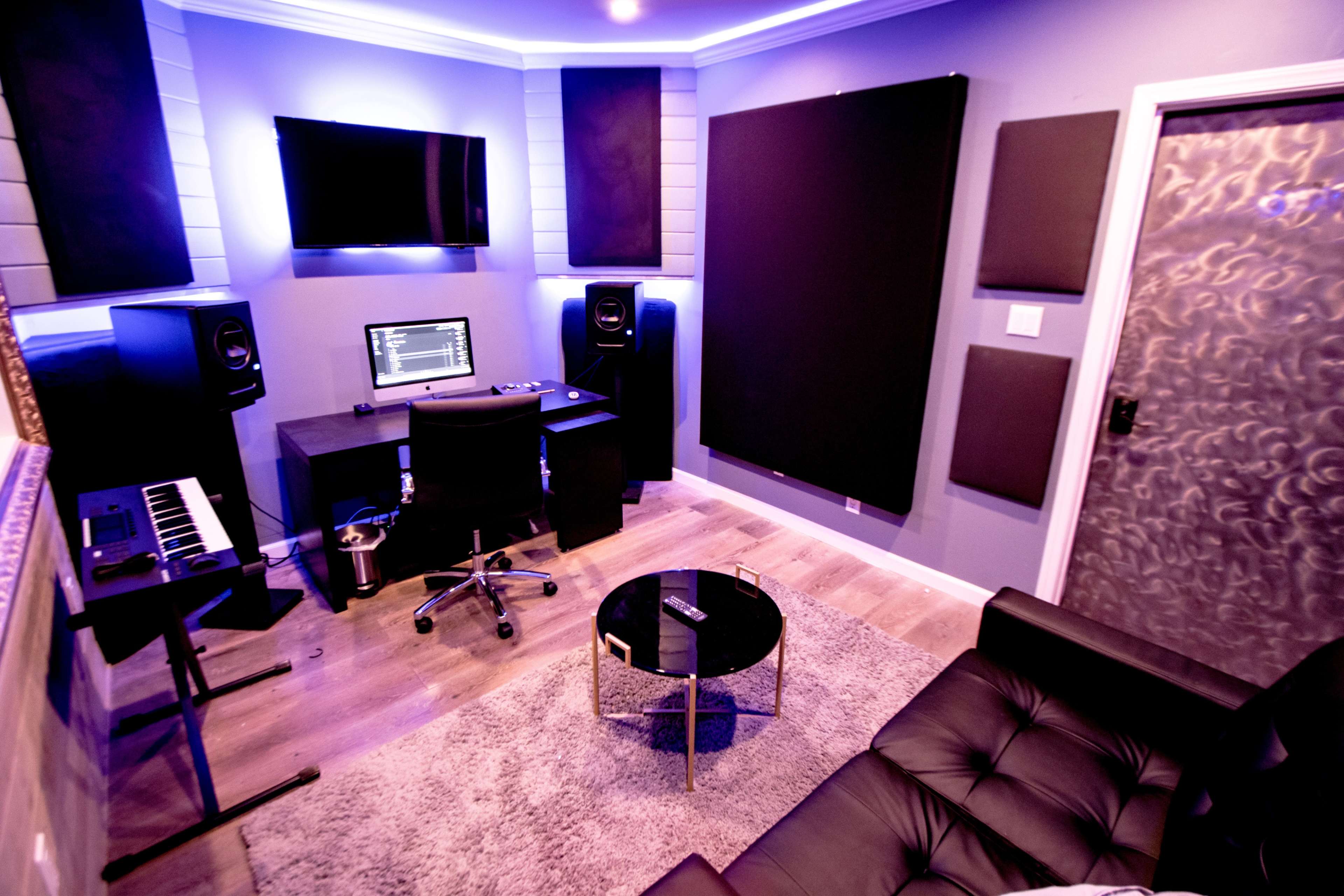 A small recording studio featuring a desk with a computer and speakers, a keyboard, soundproofing panels on the walls, and a seating area with a black couch and coffee table.