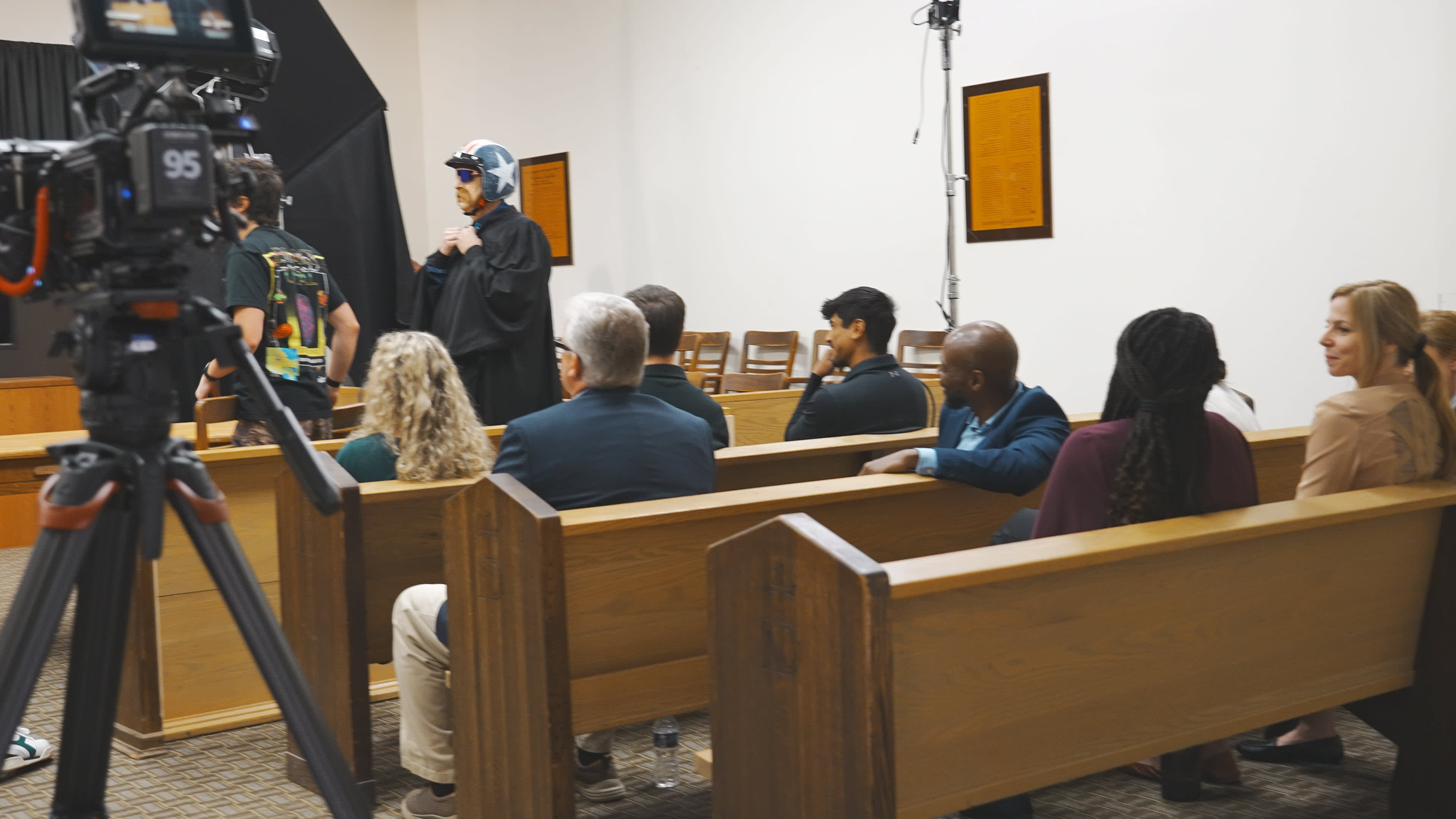 A group of people sits in wooden pews in a courtroom setting, while a person in a costume stands near a camera and a lighting setup.