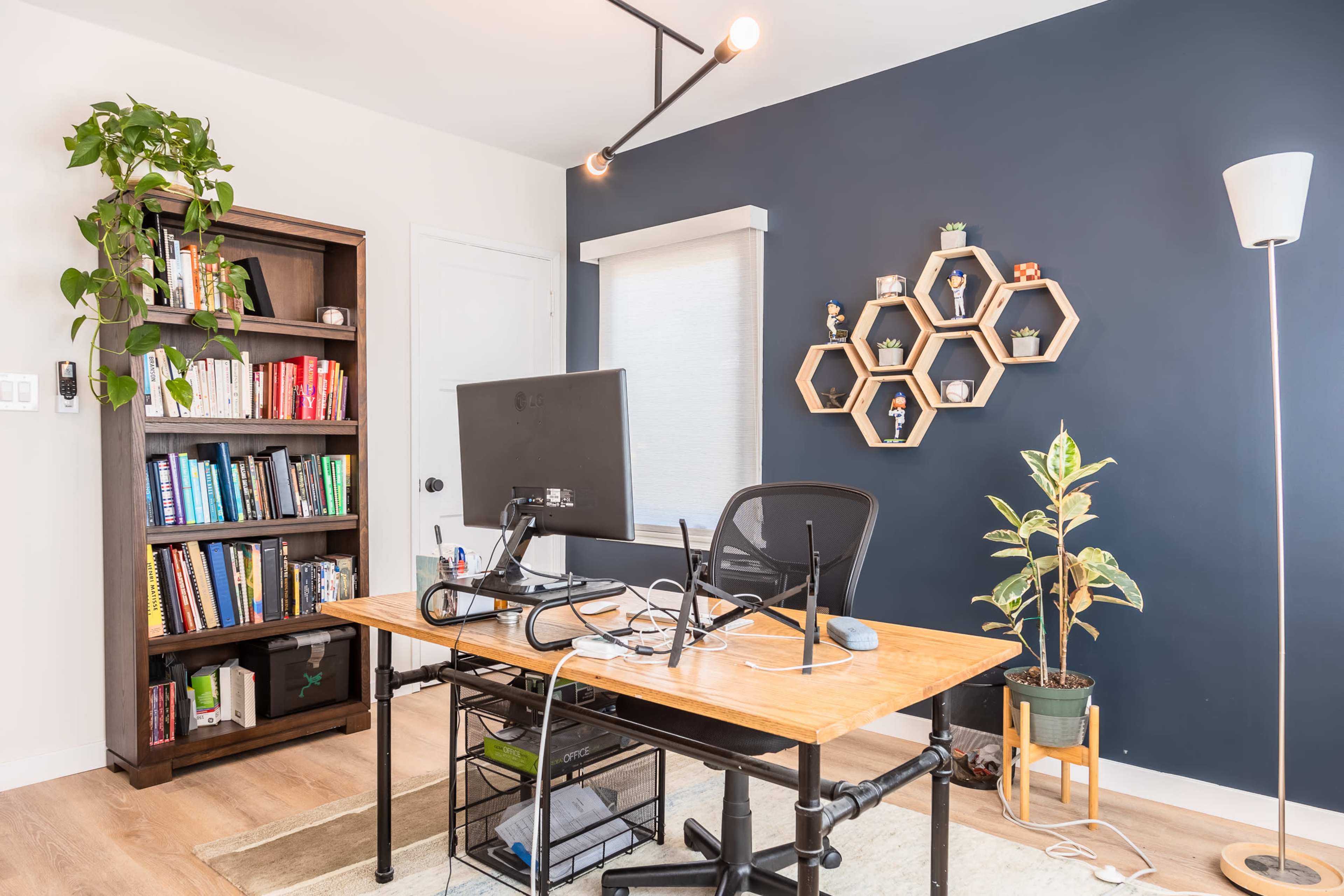 A modern home office features a desk with a computer, a bookshelf filled with books, and decorative hexagonal shelves on the wall.