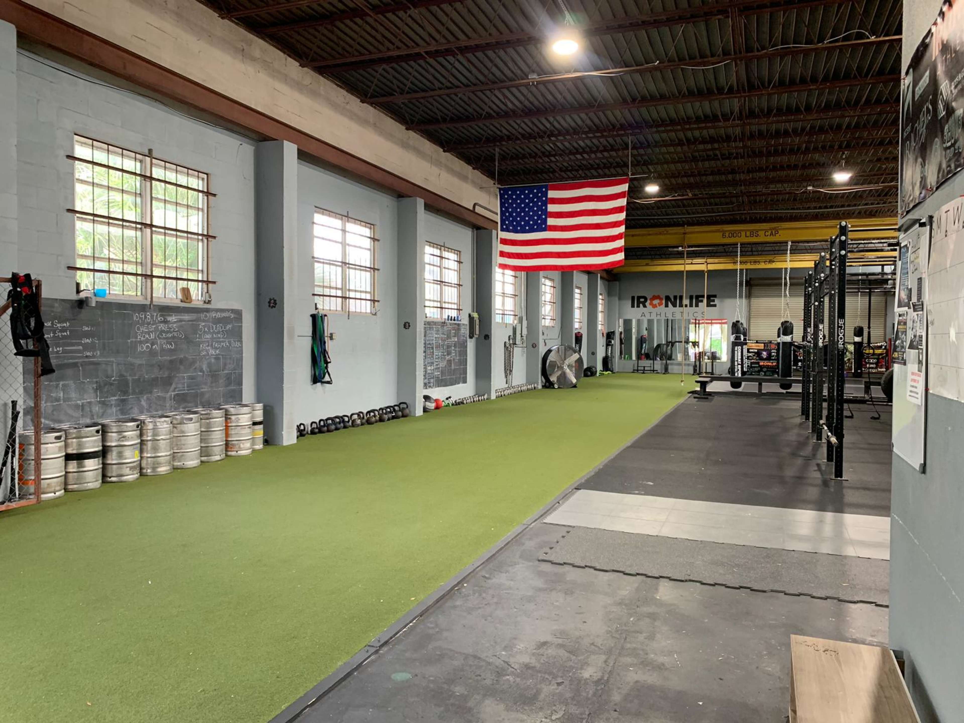 A spacious gym interior featuring green turf flooring, weightlifting equipment, and an American flag hanging on the wall.
