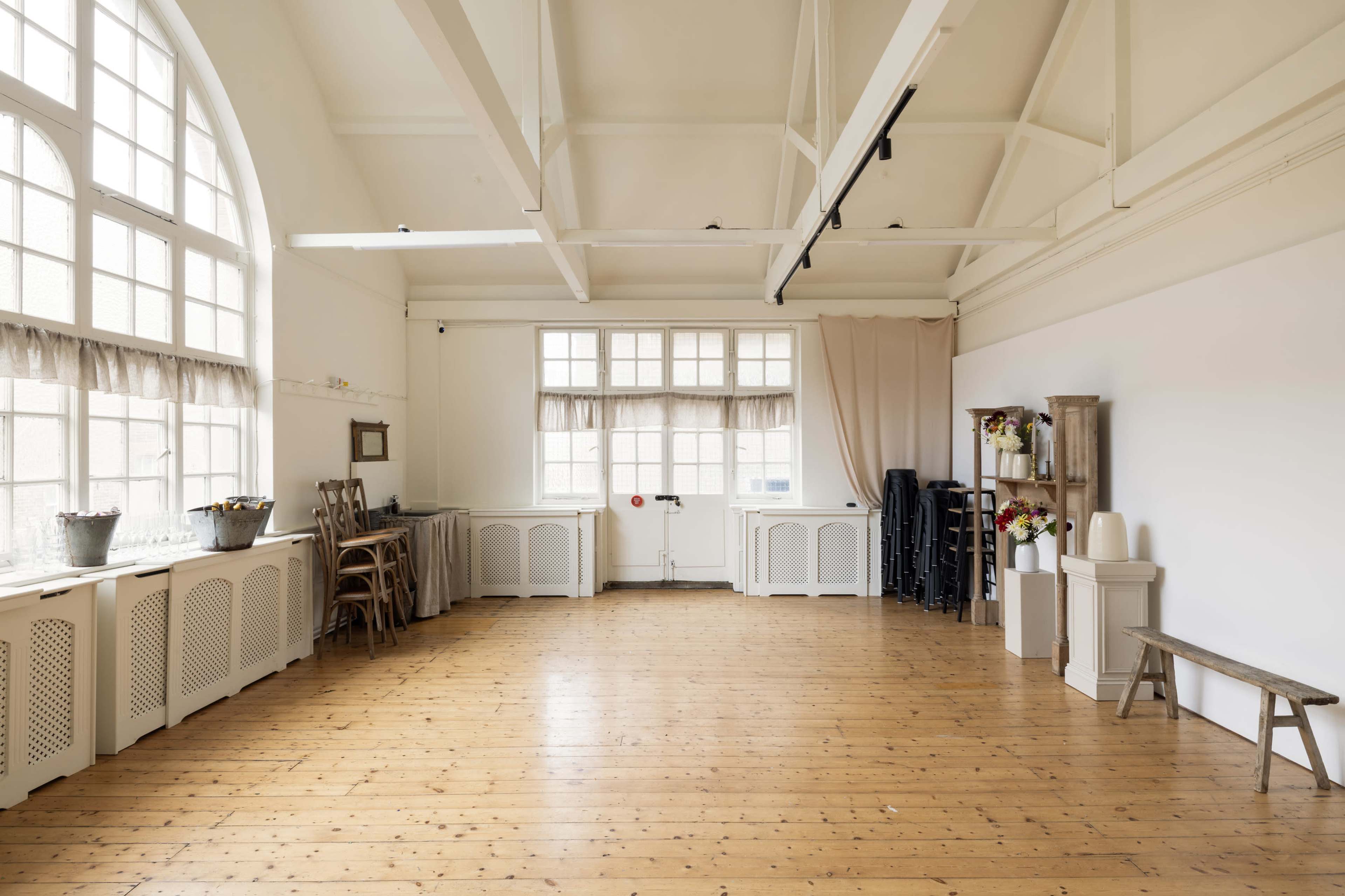 The image shows a spacious interior room with wooden flooring, large windows, and minimal furnishings, including a table, chairs, and cabinets along the walls.