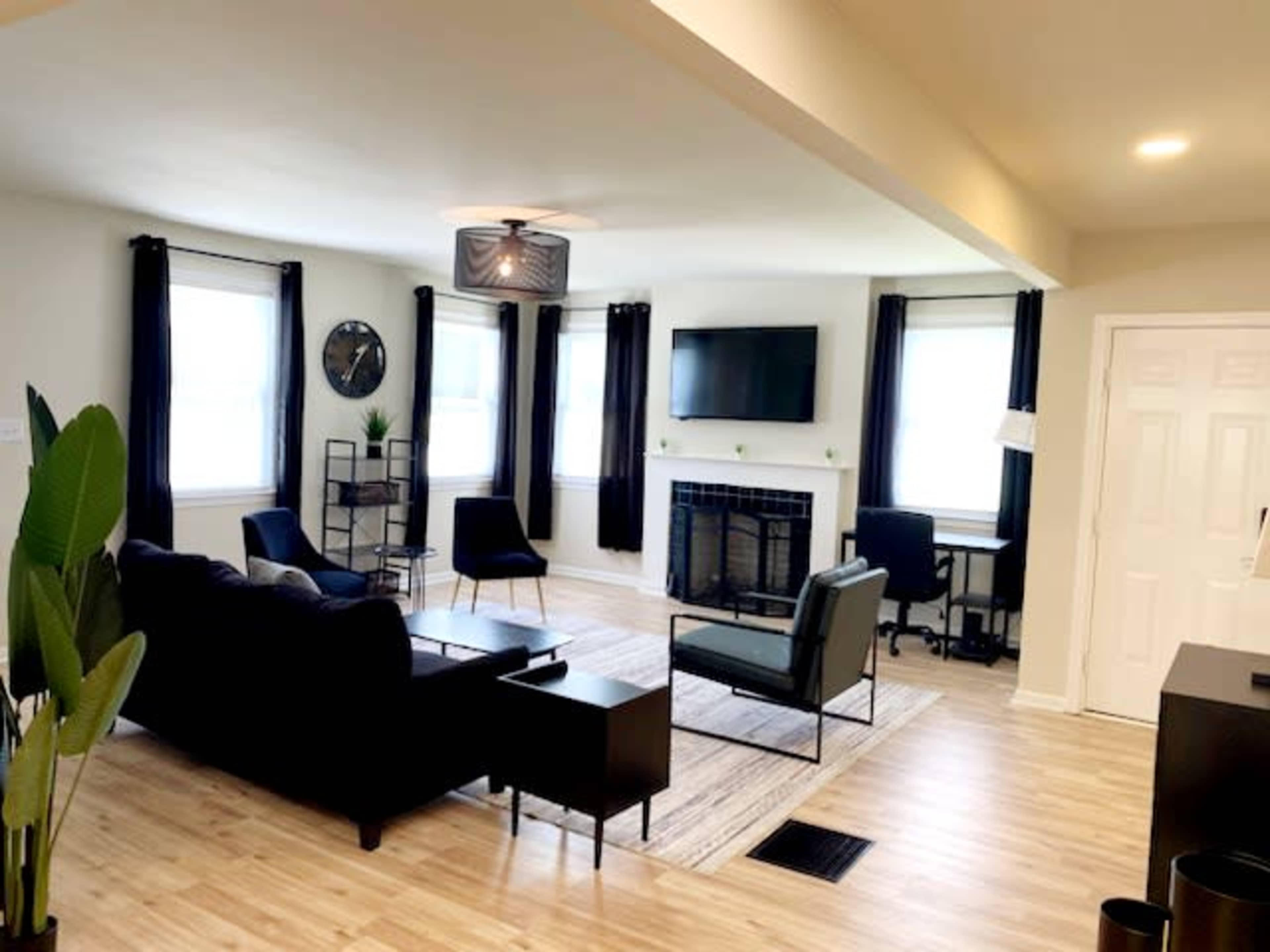 The room features a modern living space with a black sofa, two chairs, a coffee table, and a desk, illuminated by natural light streaming through large windows.