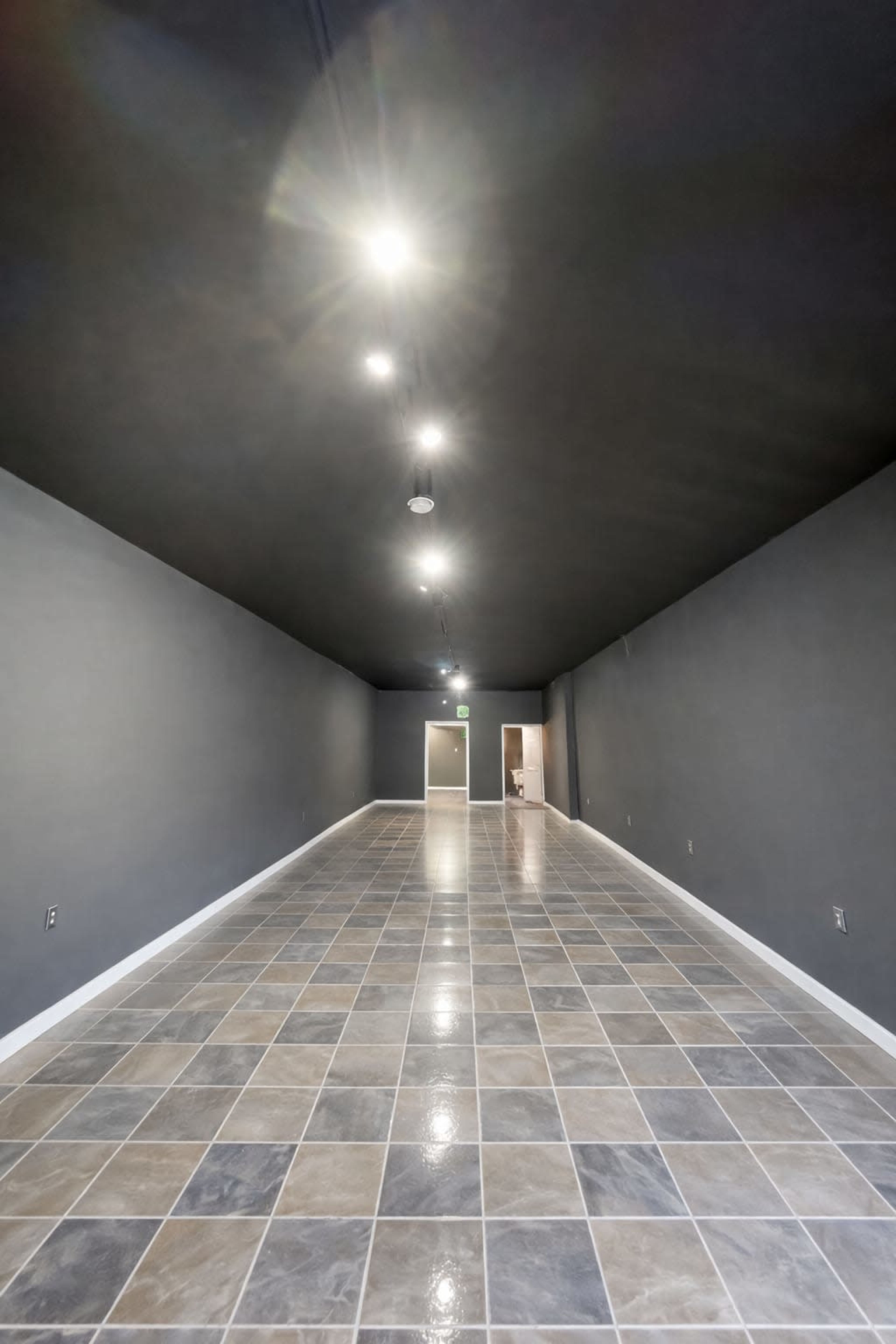 The image depicts a spacious, empty room with a long, tiled floor and gray walls, illuminated by ceiling lights.