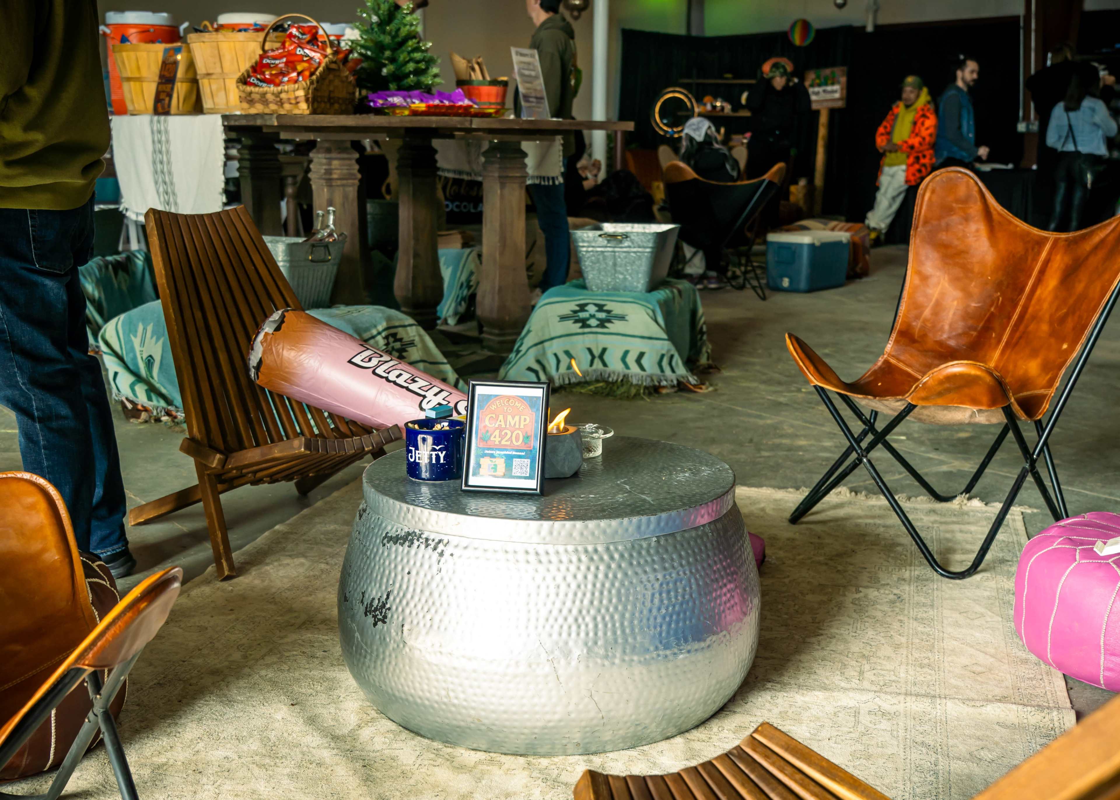 The image shows a cozy indoor space with various seating options, including wooden chairs and metal stools, arranged around a silver, round table that holds a small digital sign.