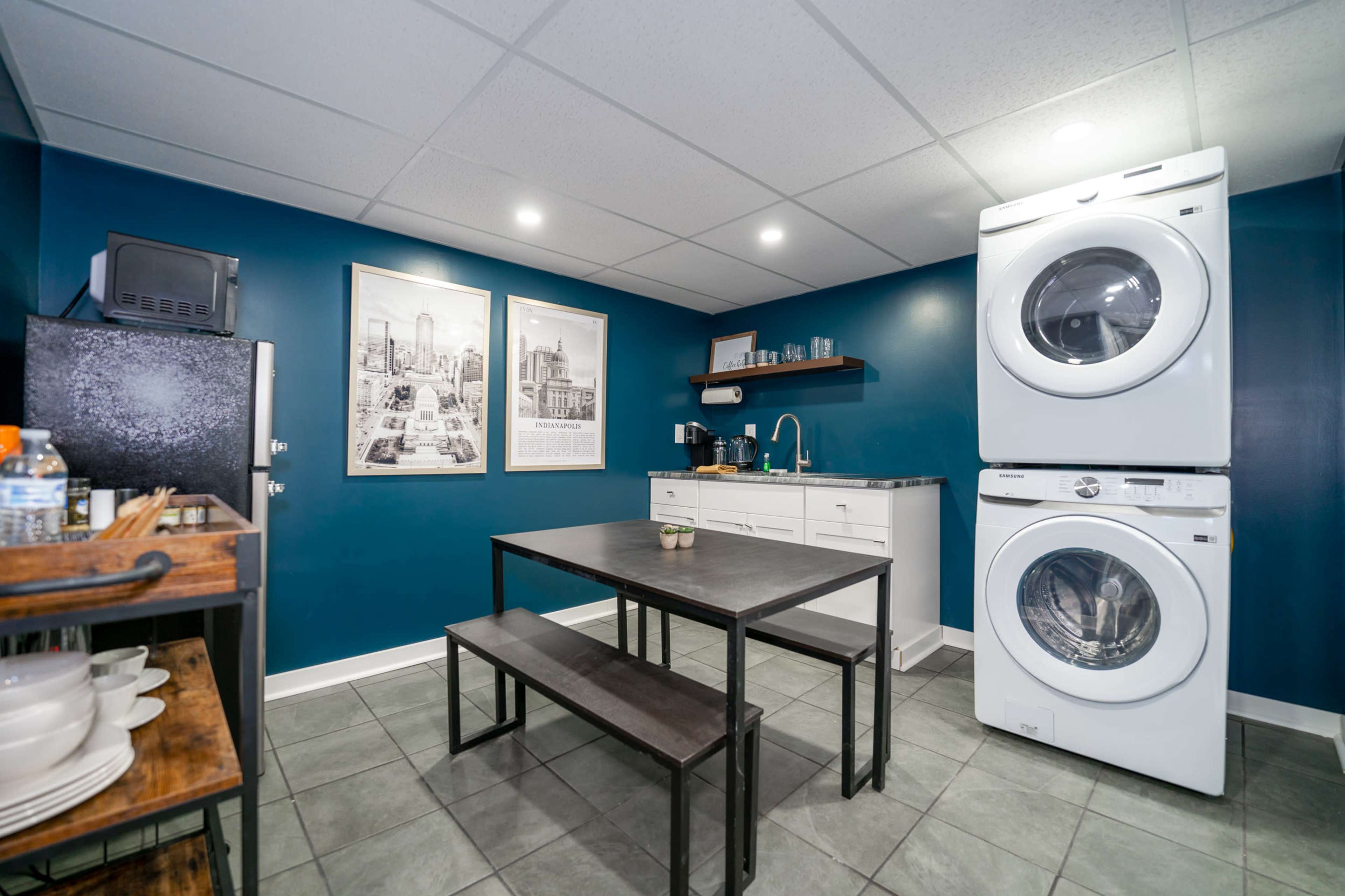 A modern kitchen space features a dark blue wall, a table with benches, and stacked washer and dryer units.