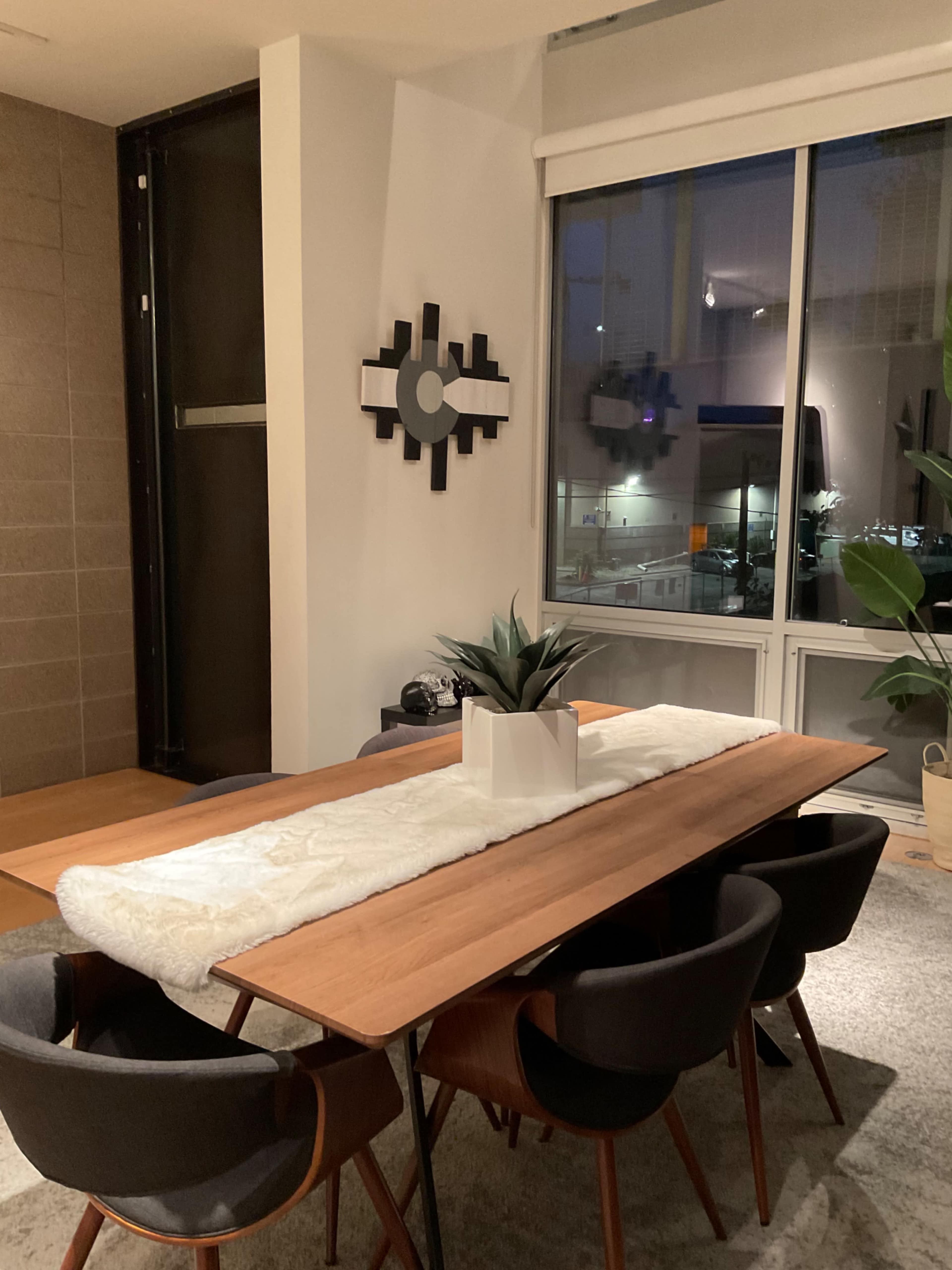 A modern dining area features a wooden table with a white runner and a succulent centerpiece, surrounded by black upholstered chairs, illuminated by evening light through a large window.