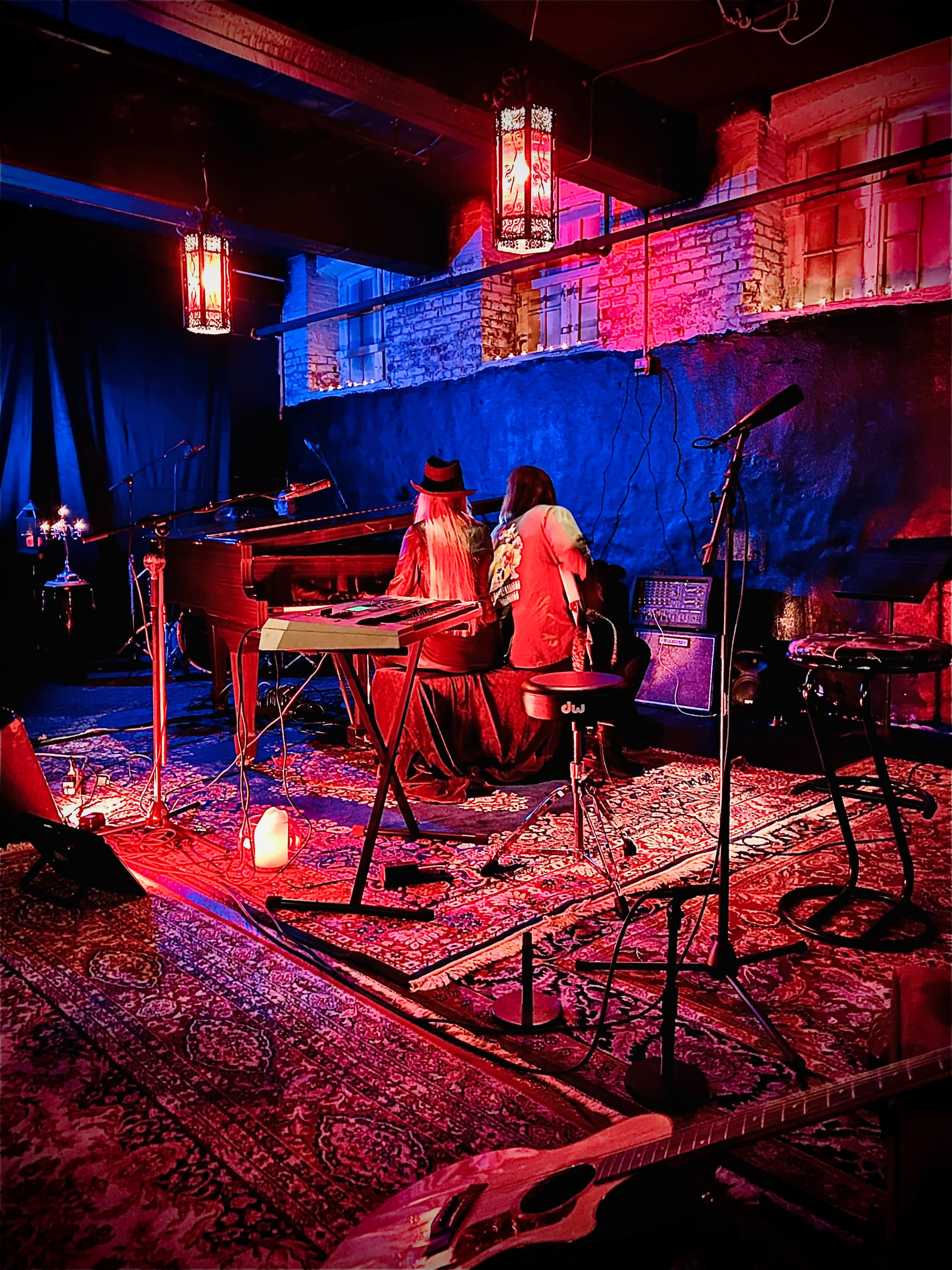 Two musicians sit at a piano and guitar in a dimly lit venue, surrounded by colorful lights and decorative lanterns.