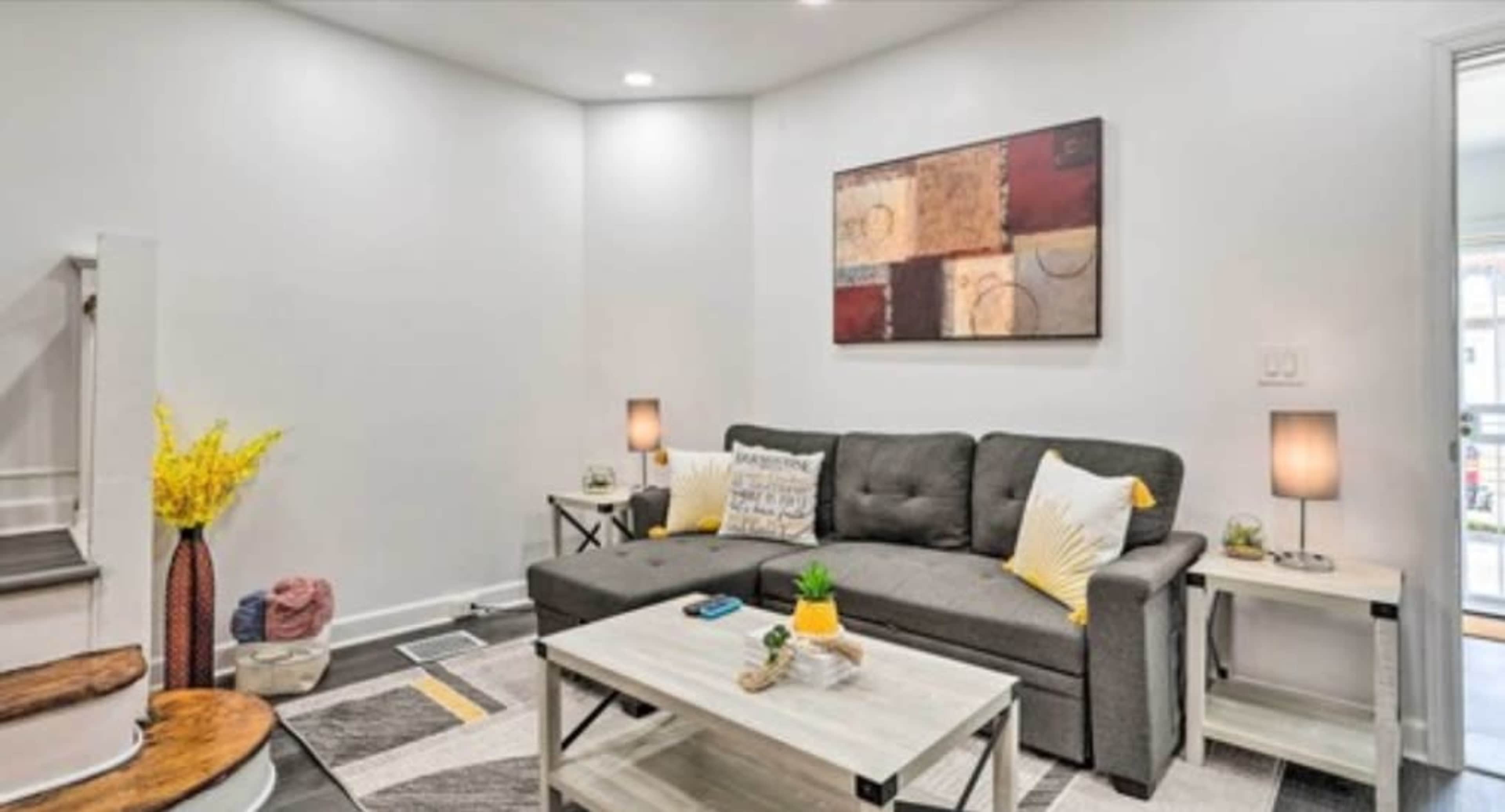 A modern living room features a gray sofa, two side tables with lamps, and a coffee table, set against white walls and decorated with a colorful abstract painting.