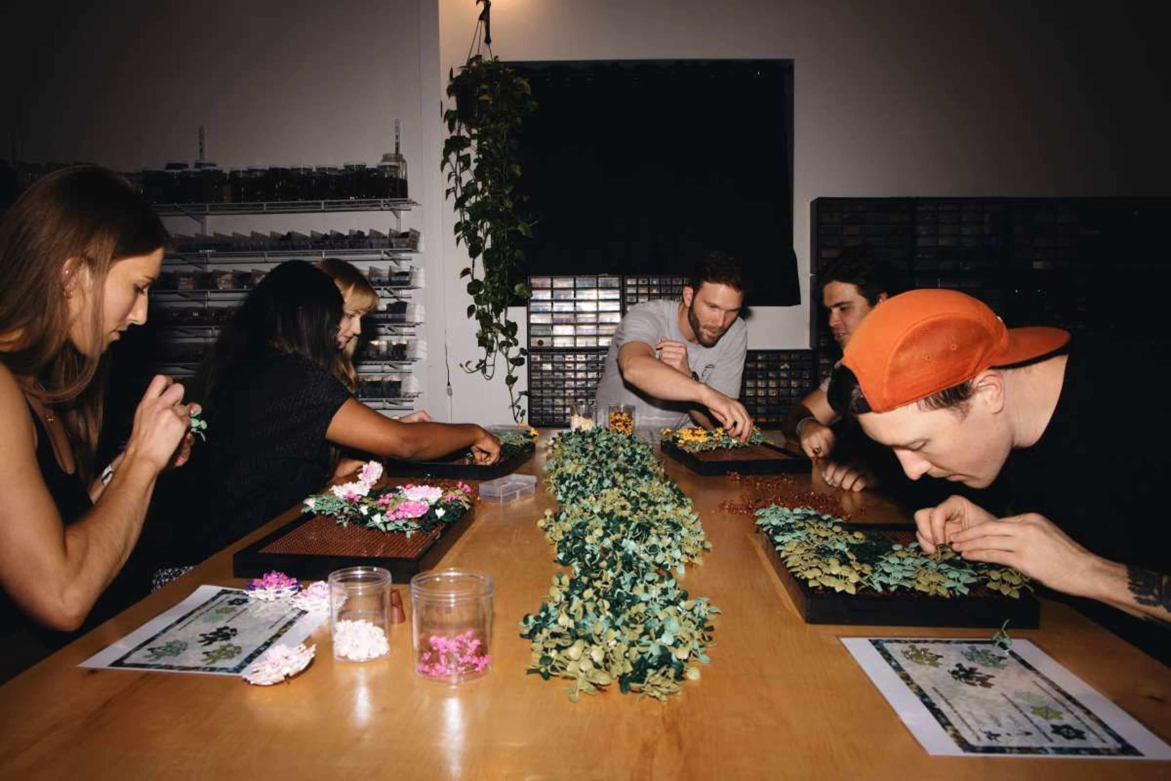 A group of six people is seated at a table, arranging various flower and leaf decorations in a workshop setting.