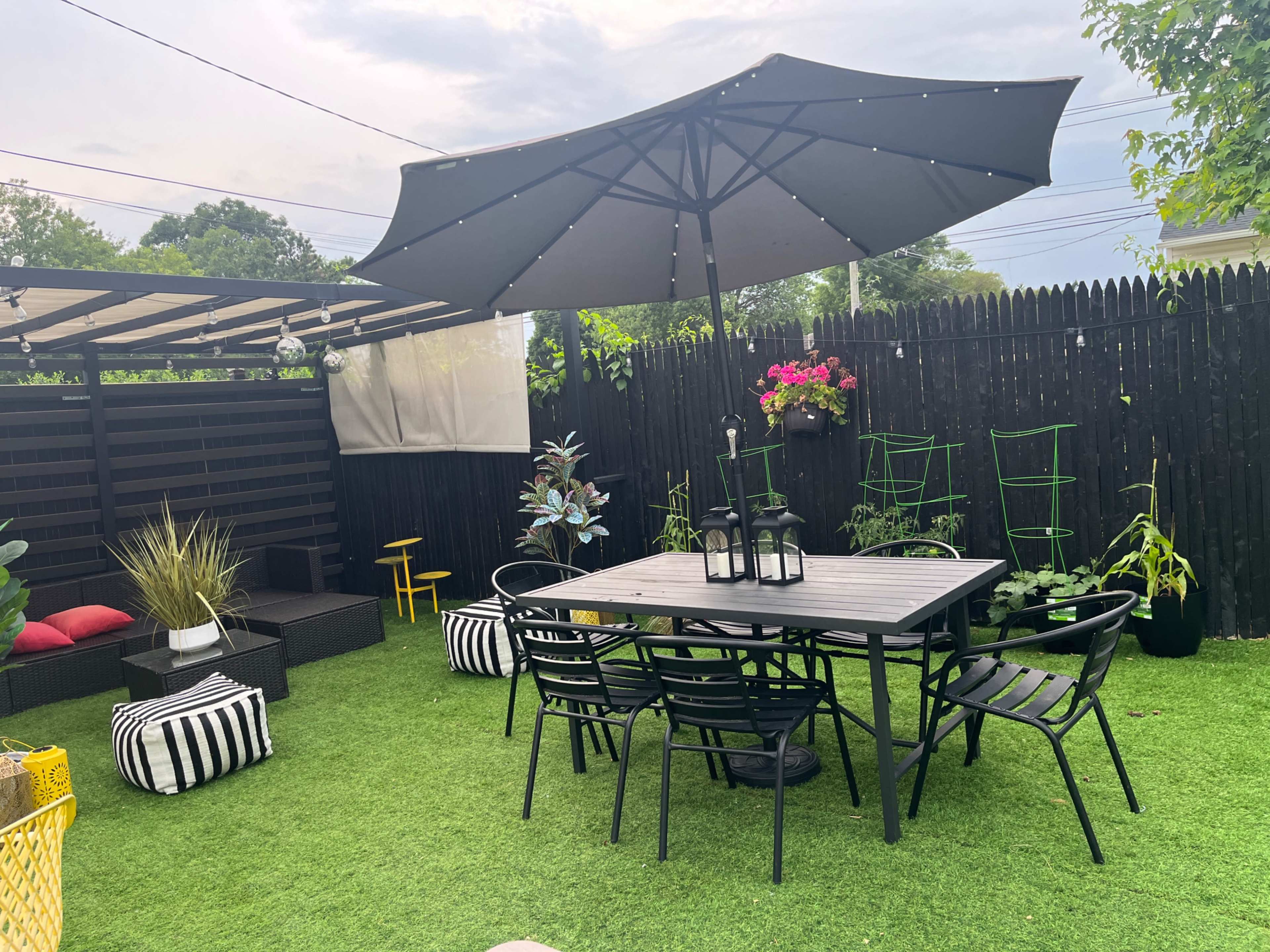 The image depicts an outdoor patio area featuring a black dining table with chairs, a large umbrella, decorative plants, and a lawn covered in artificial grass.
