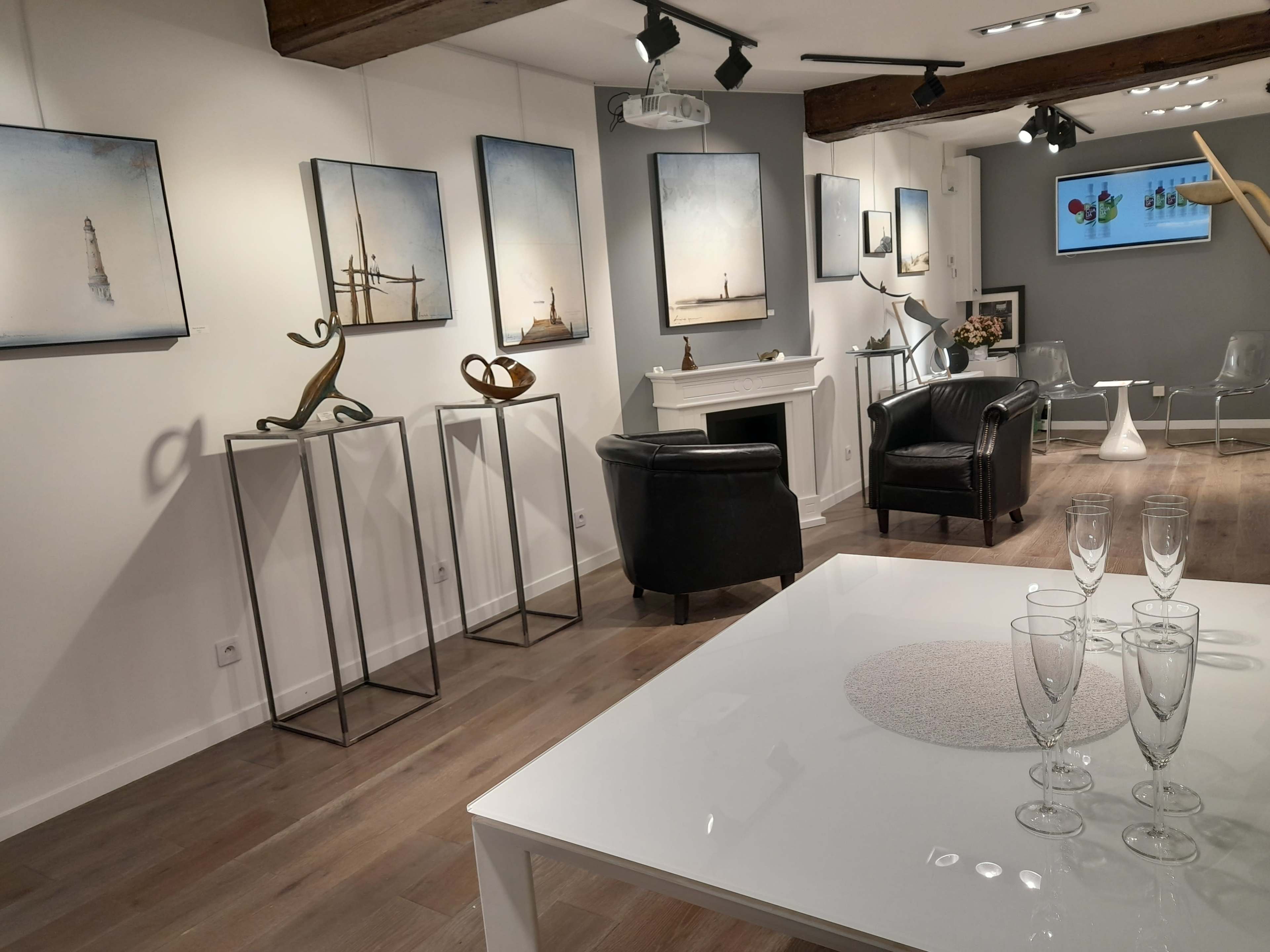 A modern art gallery featuring abstract sculptures and framed artworks on white walls, with a sleek white table set with champagne flutes in the foreground.