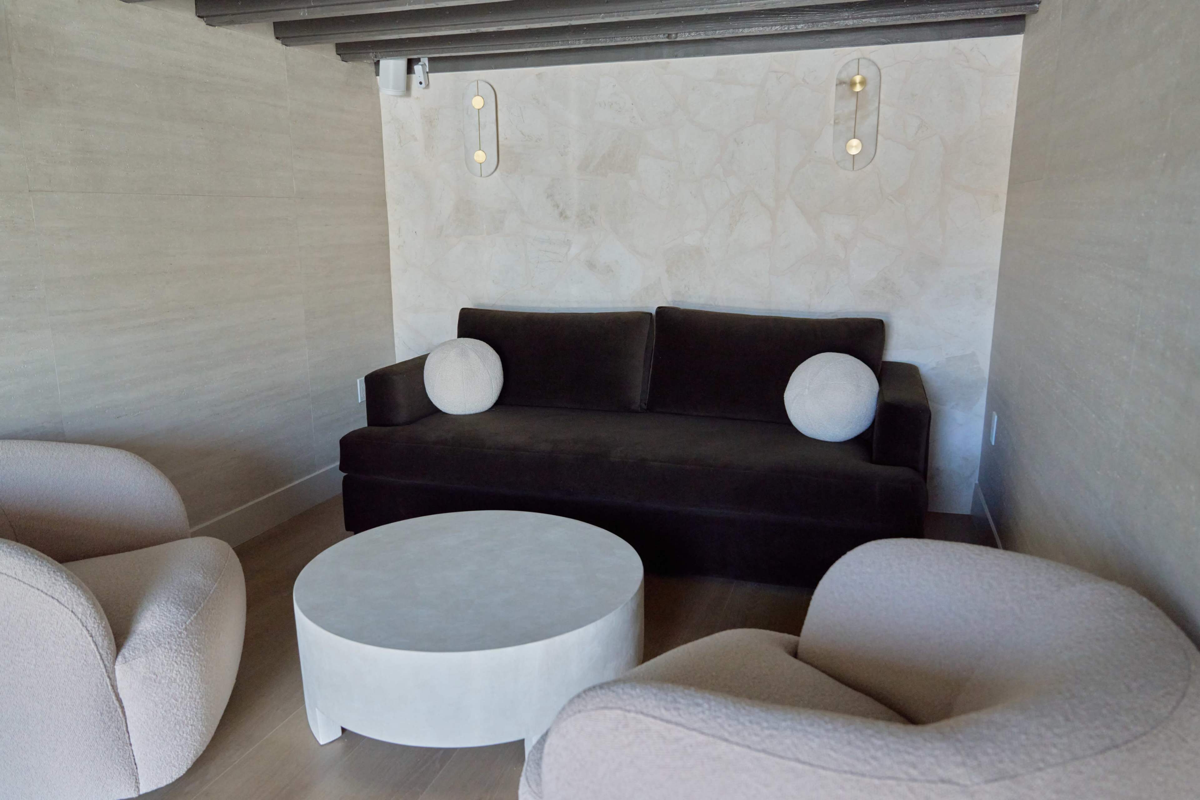 The image shows a minimalist interior design featuring a dark sofa with two round cushions, a round coffee table, and two light-colored armchairs in a softly lit room.