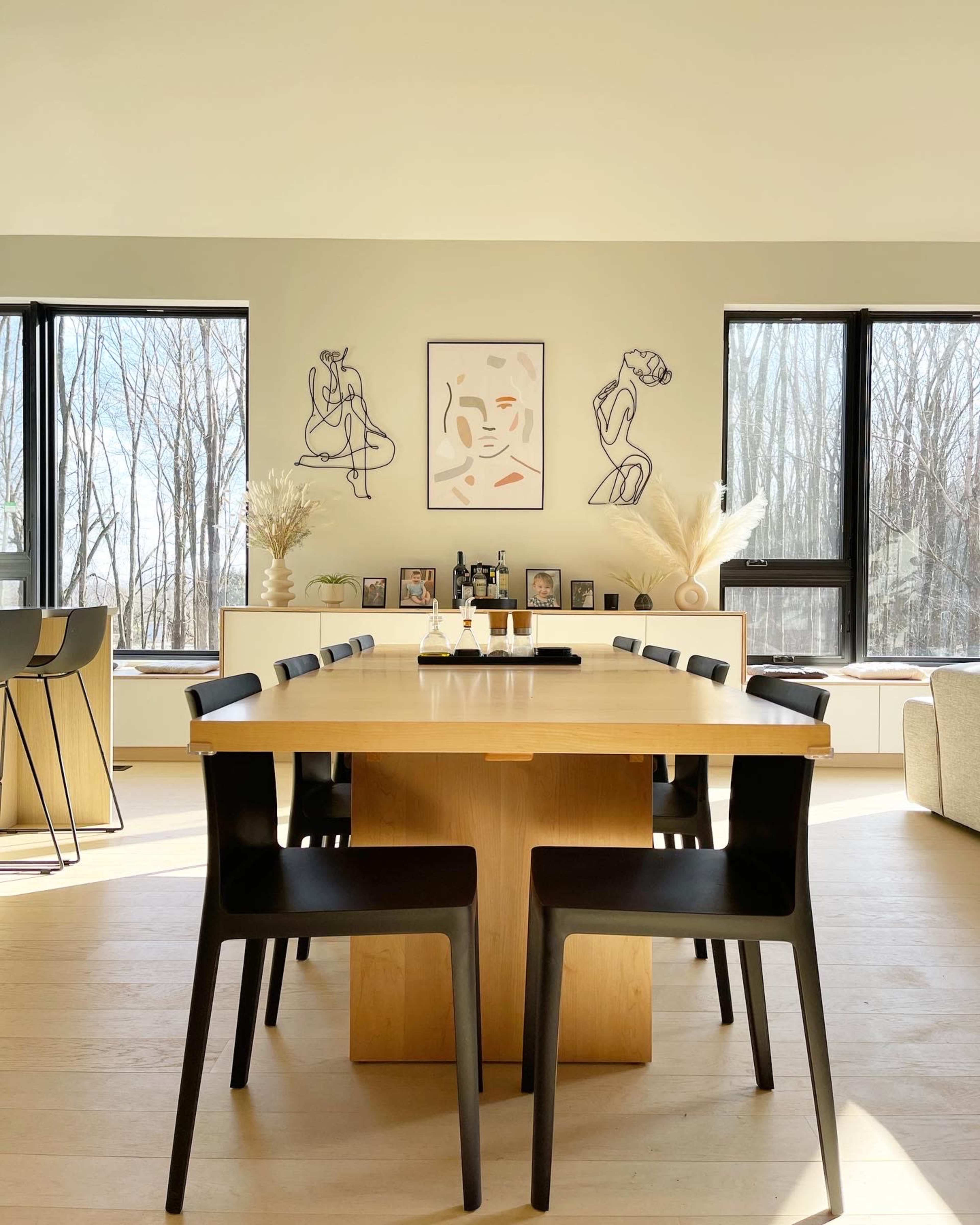 A modern dining room features a large wooden table surrounded by black chairs, with art on the walls and large windows overlooking a wooded area.