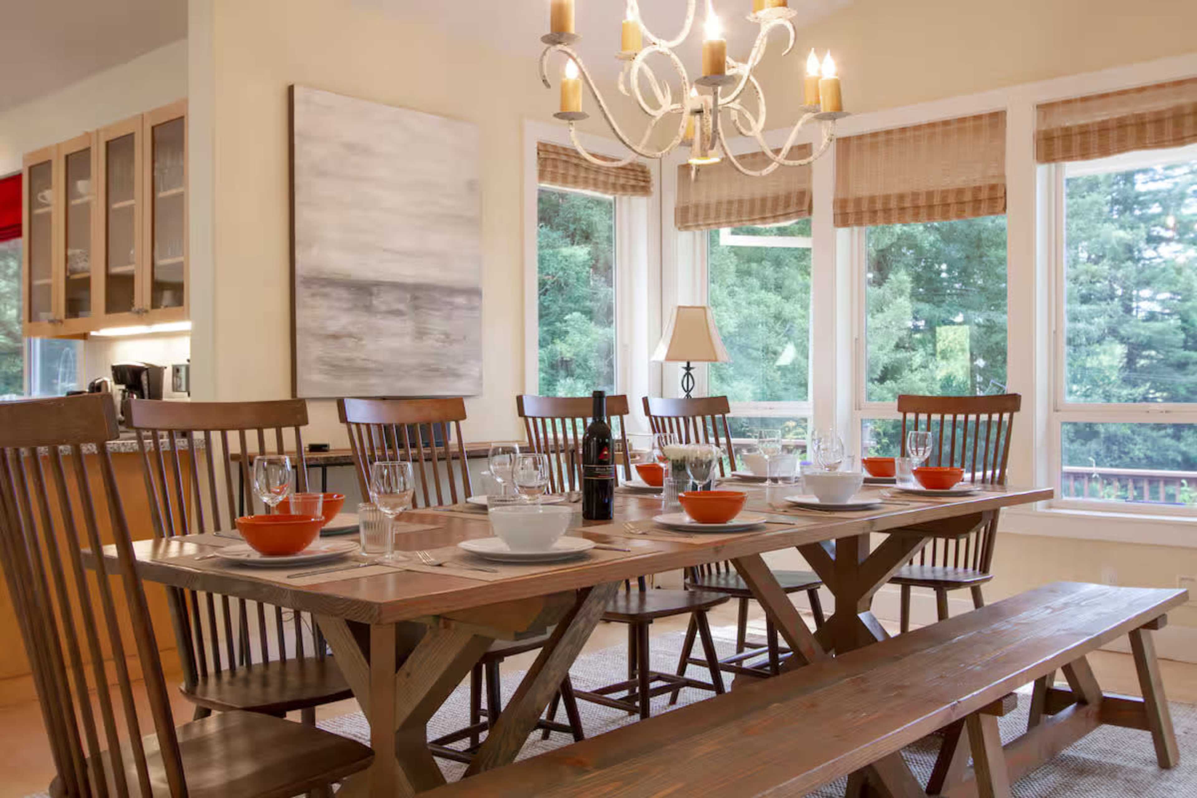 A dining room features a long wooden table set for a meal, surrounded by matching chairs and illuminated by a chandelier, with large windows offering a view of greenery outside.