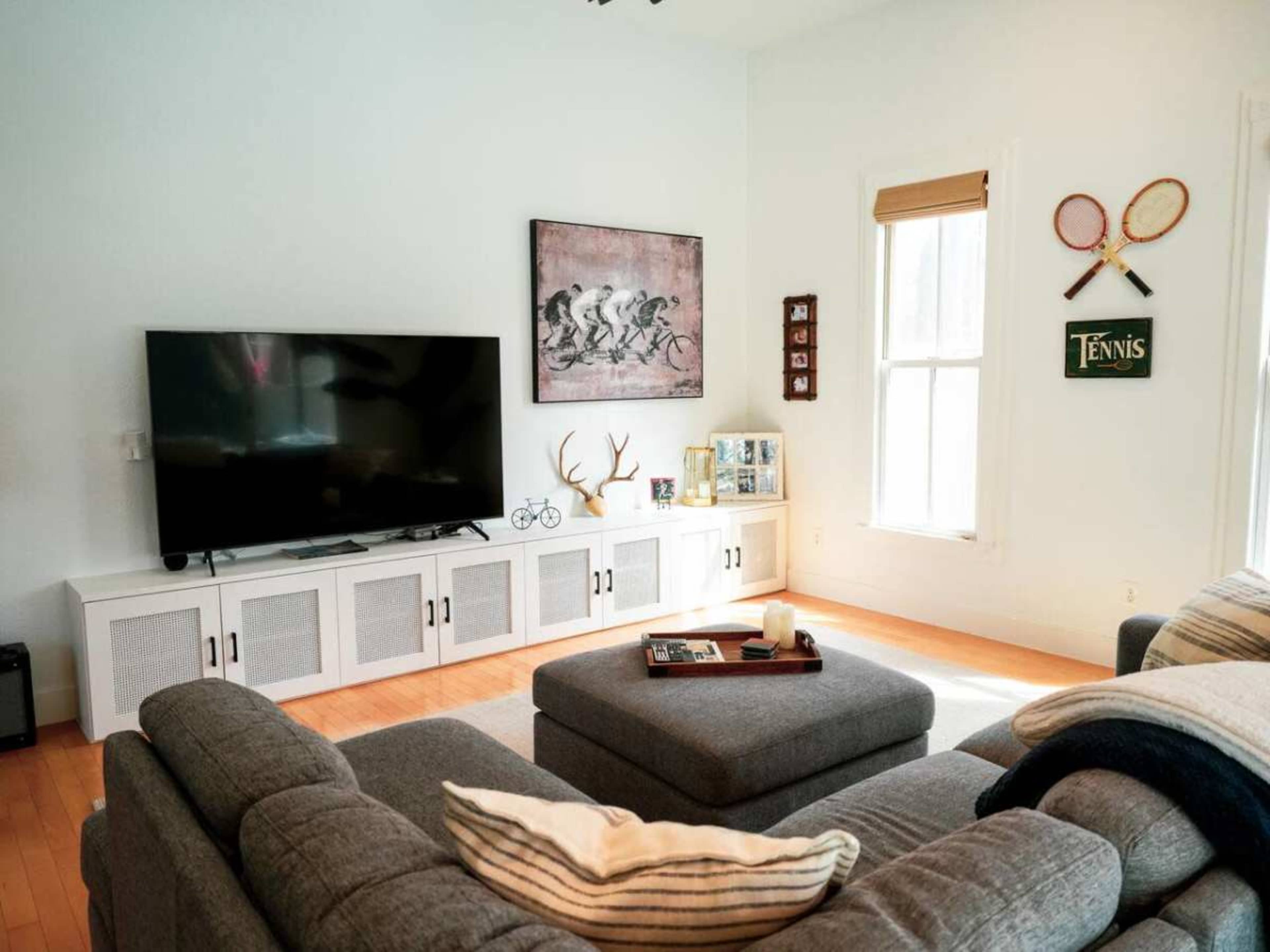 The living room features a gray sectional sofa, a large television on a white entertainment unit, and decor that includes a tennis sign and artwork on the walls.