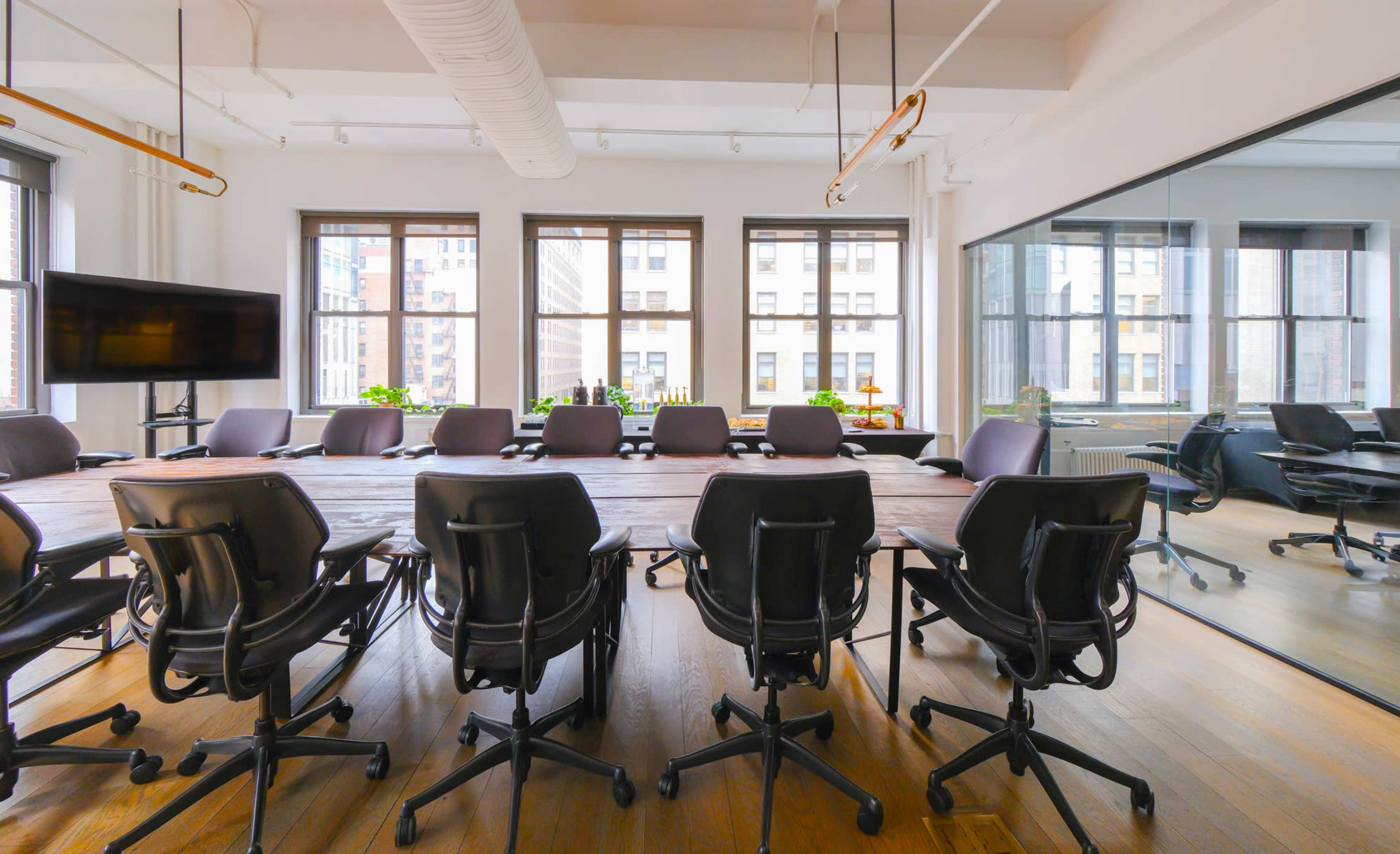 A large conference room features a long wooden table surrounded by black office chairs, with windows allowing natural light to illuminate the space.