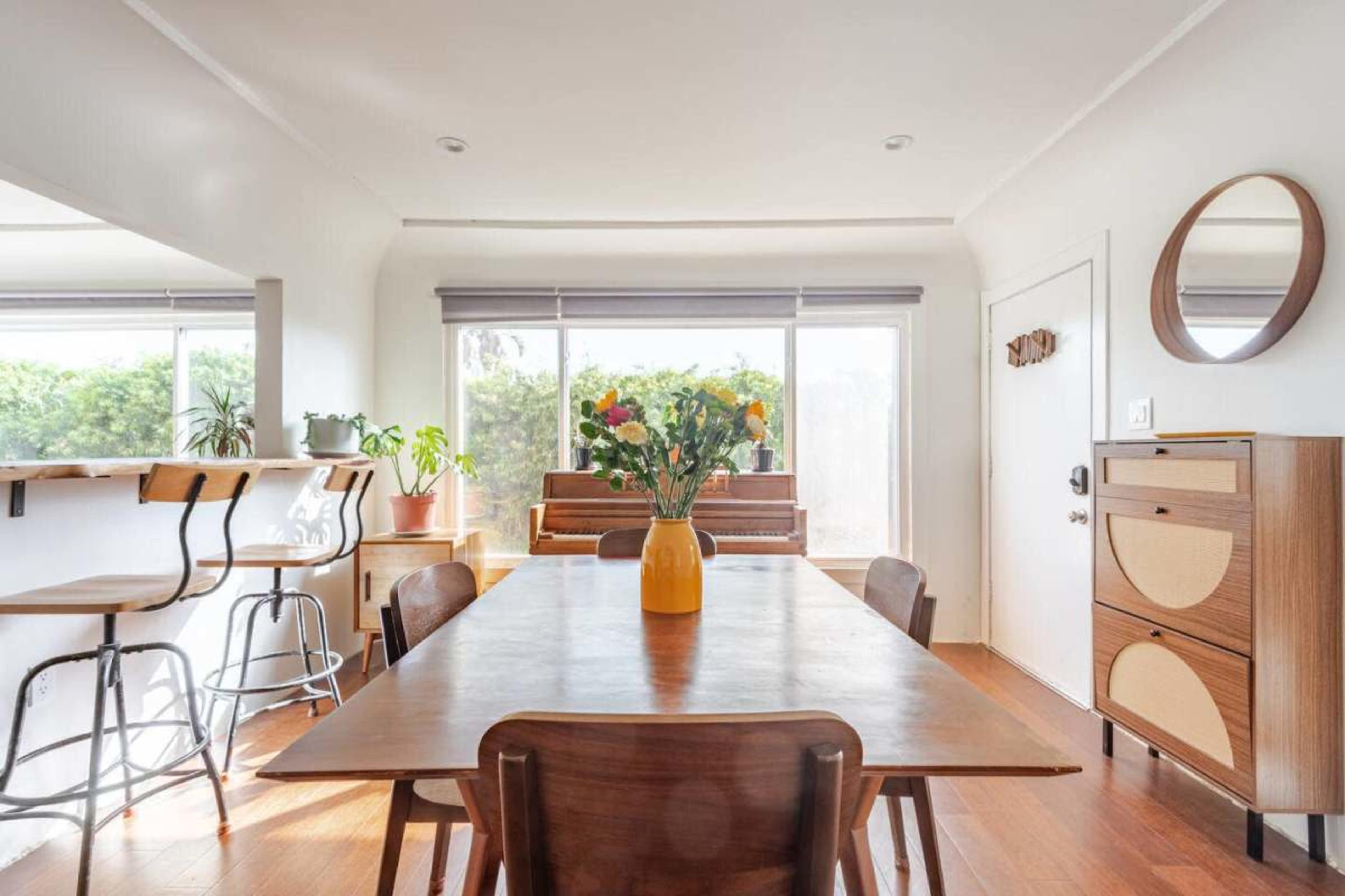 A modern dining area features a wooden table with a vase of flowers, surrounded by chairs, and a view of a piano and greenery through large windows.