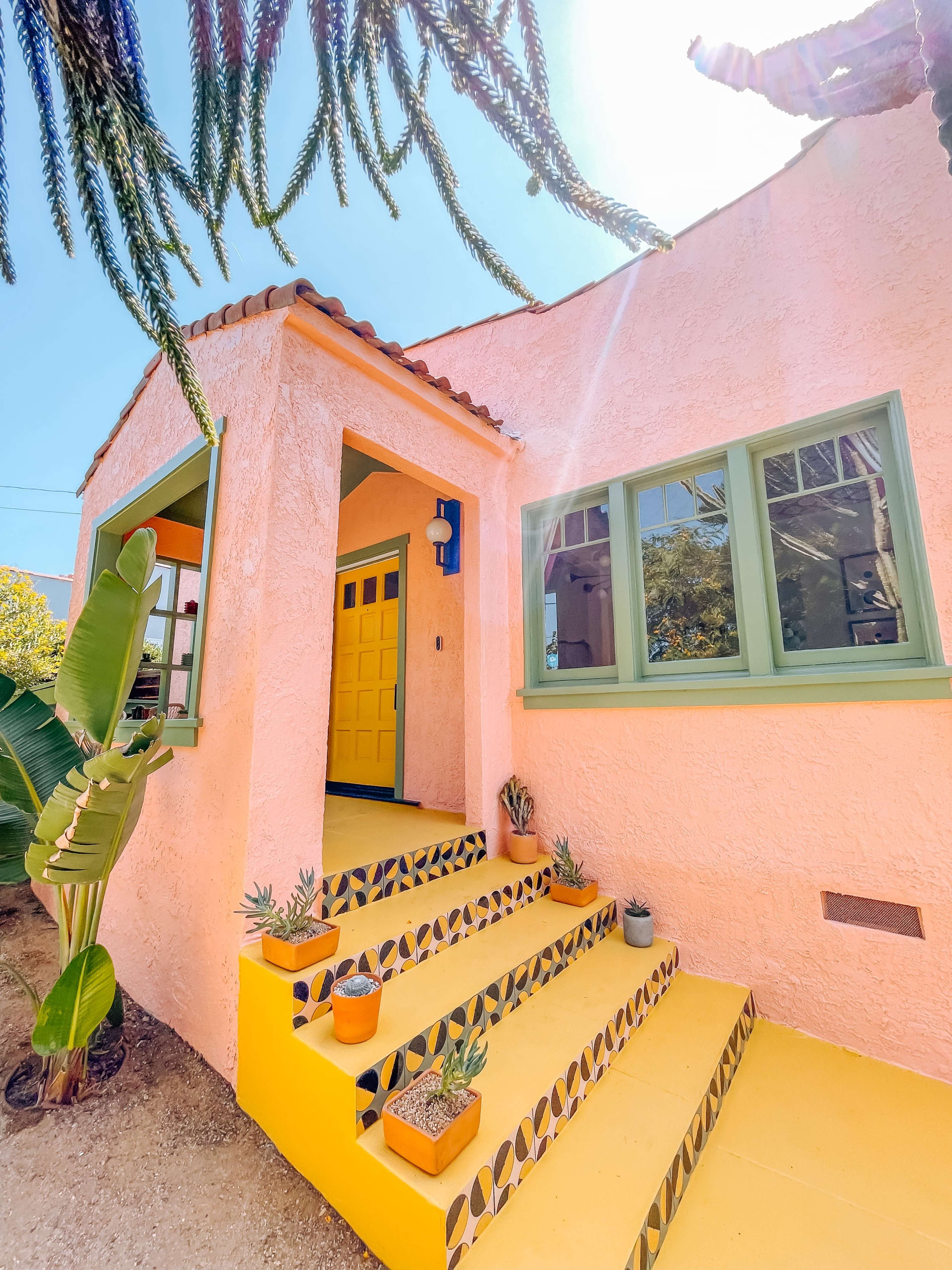 A pink house with green window frames features a yellow staircase adorned with potted plants and colorful tiles.