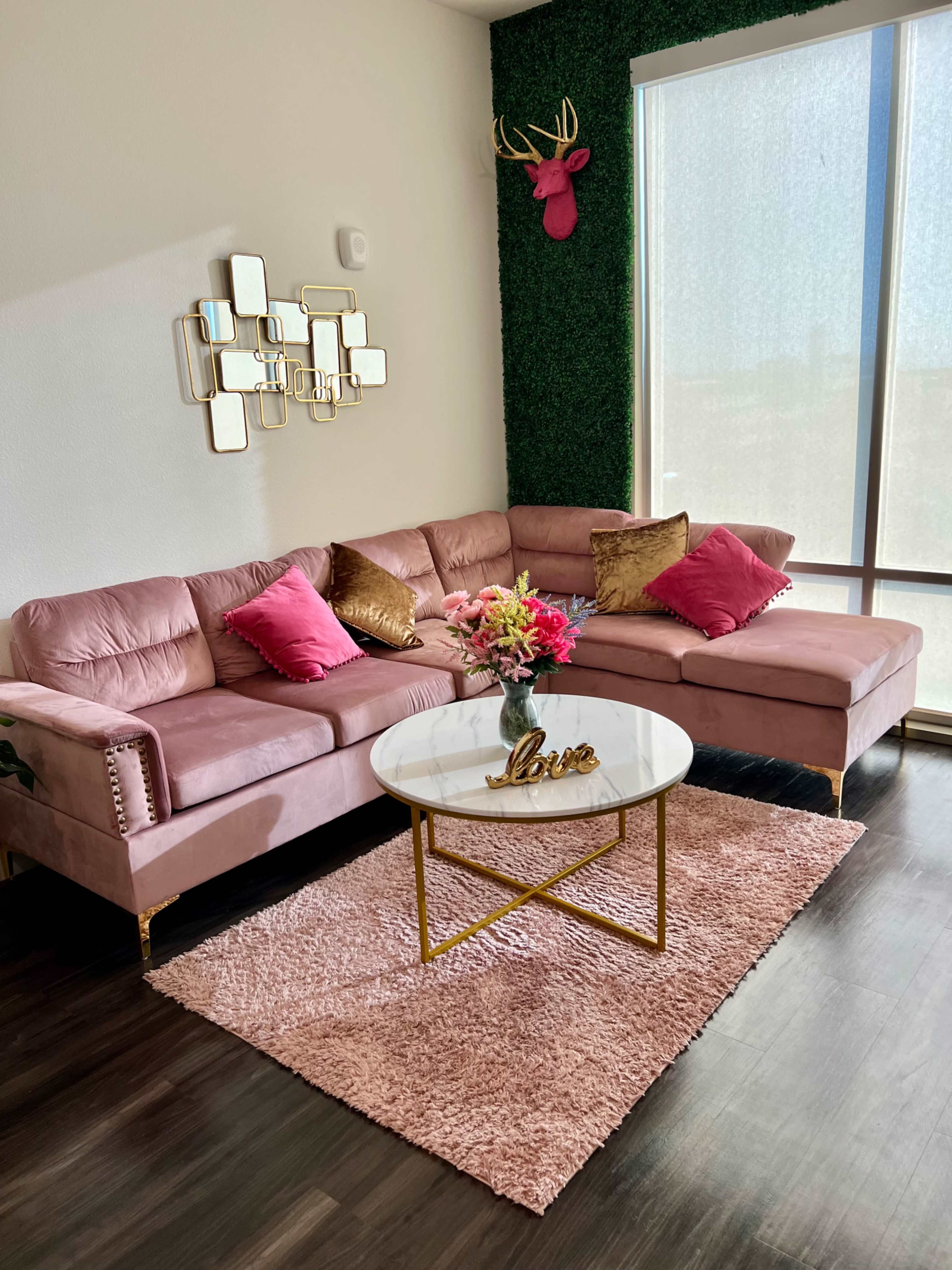 A pink sectional sofa with decorative pillows sits on a fluffy rug in a bright room featuring a green wall and large windows.