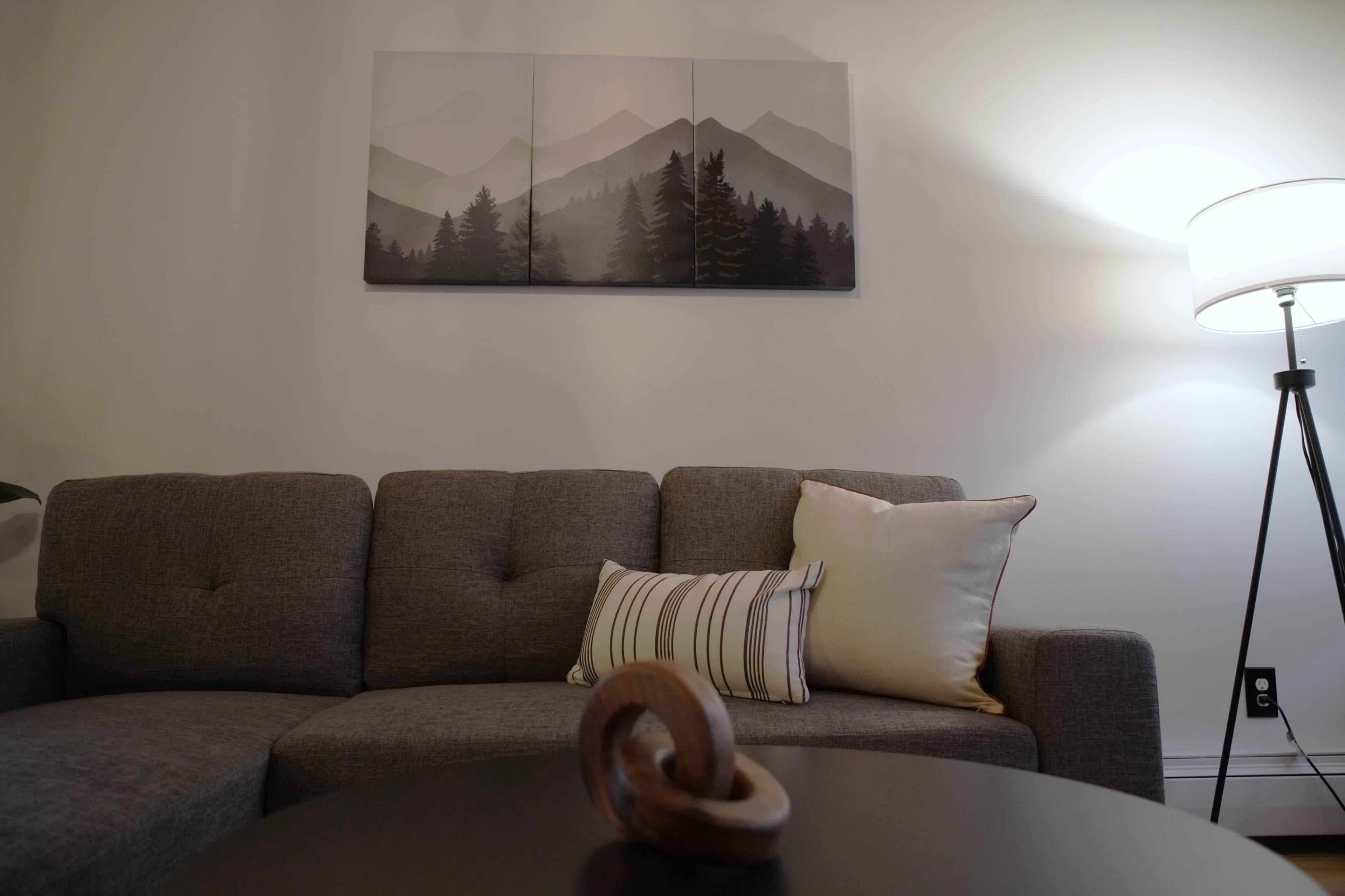 A modern living room features a gray sofa with decorative pillows, a round coffee table with a wooden sculpture, and a landscape painting of mountains on the wall.