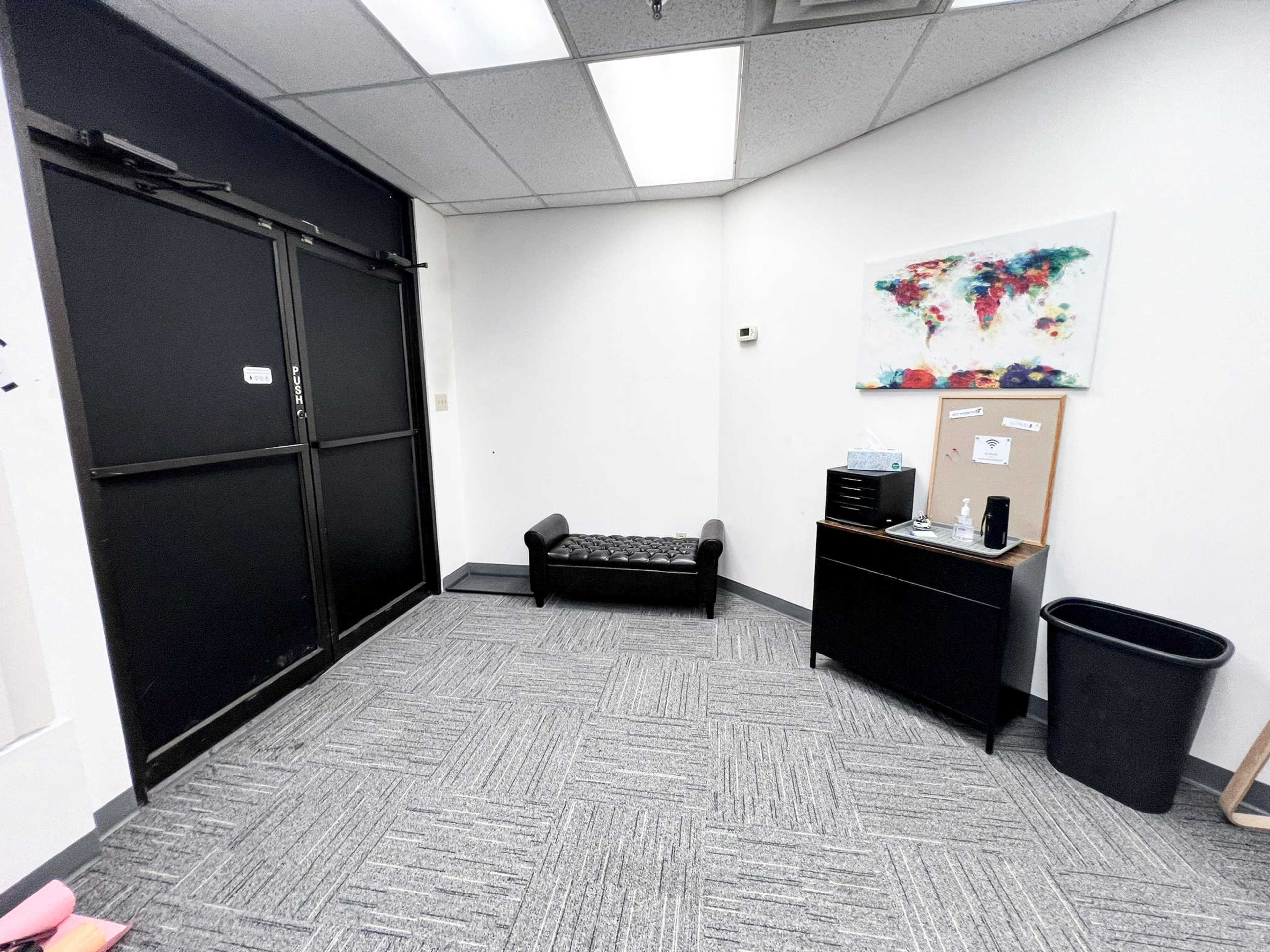 A small office space with a black double door, a bench, a dark filing cabinet, and a trash can against a white wall.