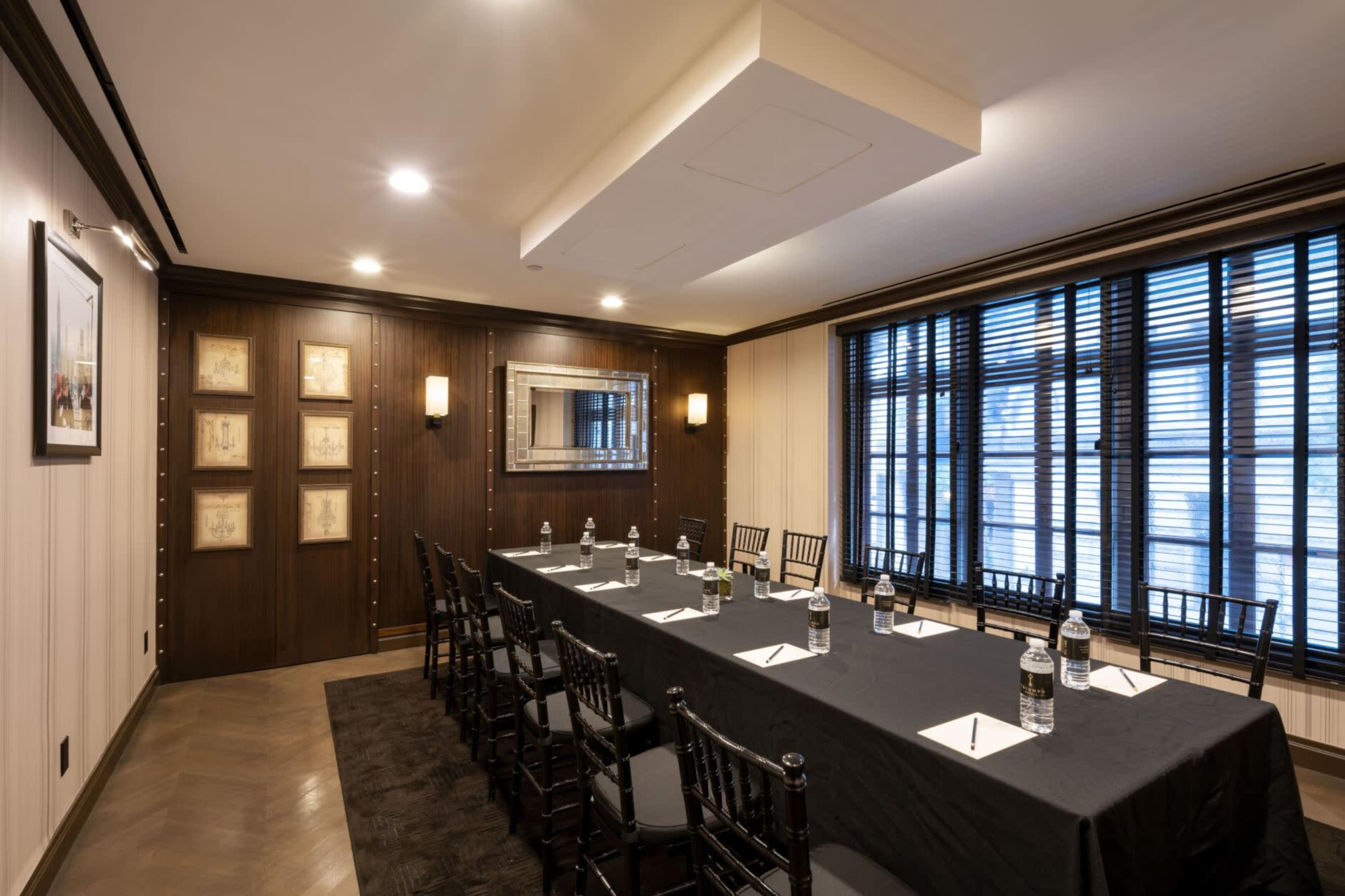 A conference room features a long rectangular table with chairs, water bottles, and notepads, set against a backdrop of wooden paneling and large windows with blinds.