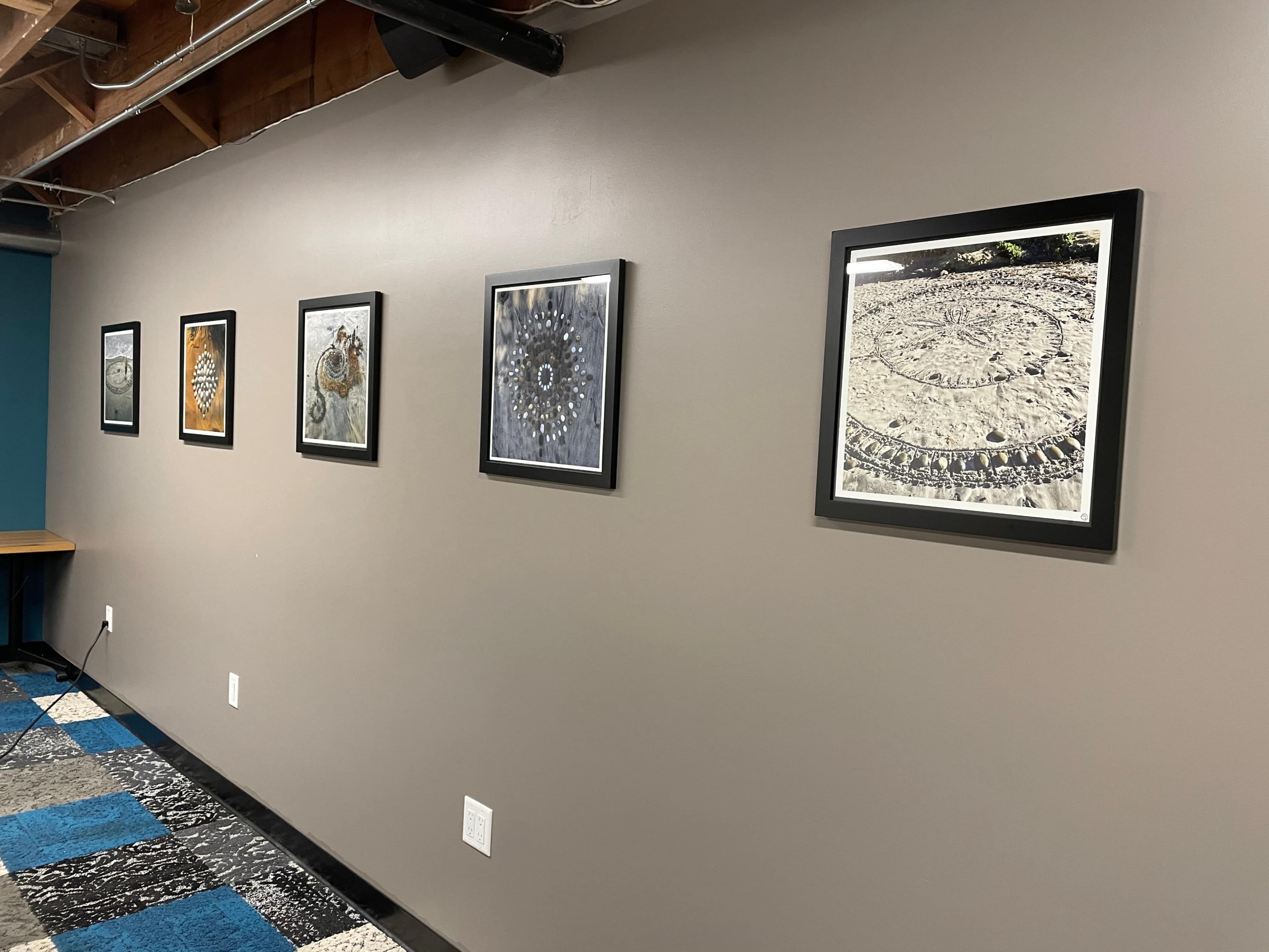 A series of framed photographs displayed on a gray wall in a basement, with a table visible to the left and a patterned floor.