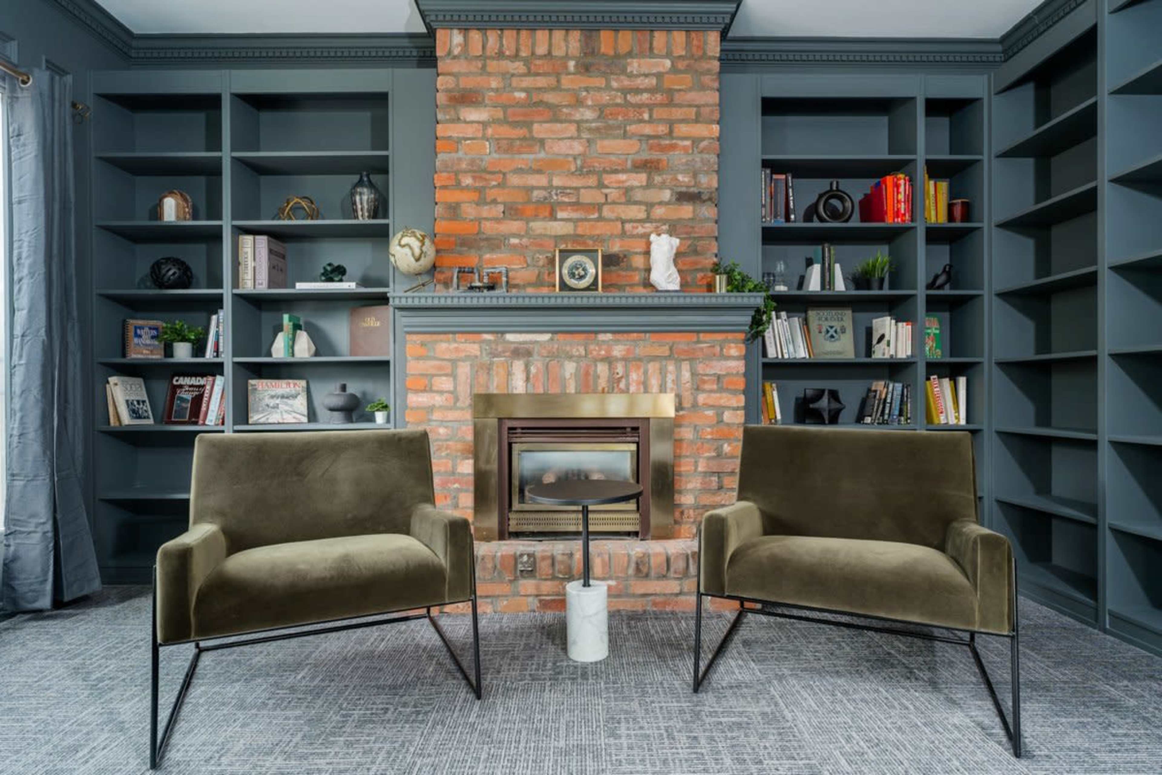 A cozy living room with two green armchairs facing a brick fireplace, surrounded by built-in shelves filled with books and decorative items.
