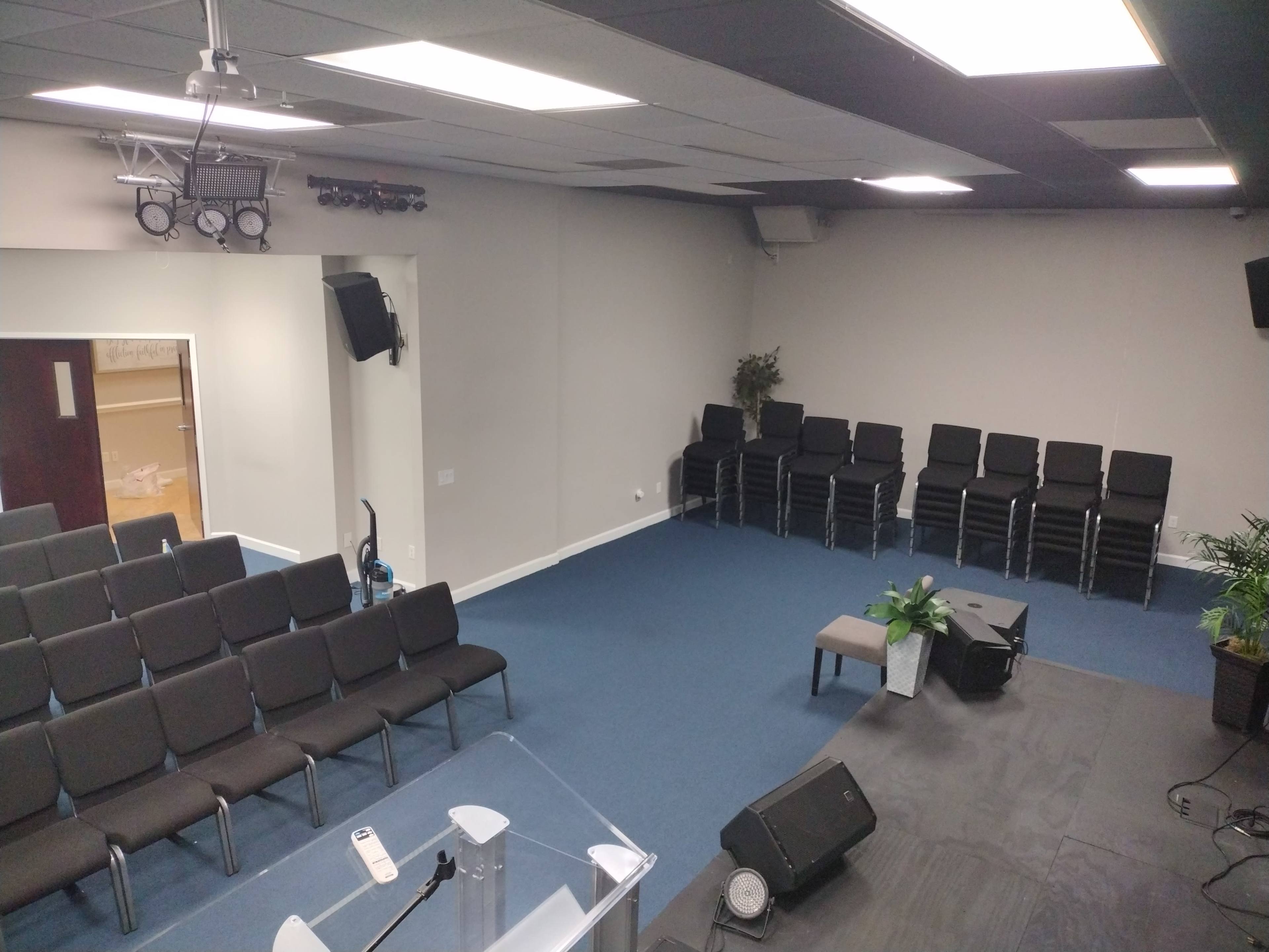 An empty indoor assembly space with rows of stacked chairs, a podium, and a small stage area.