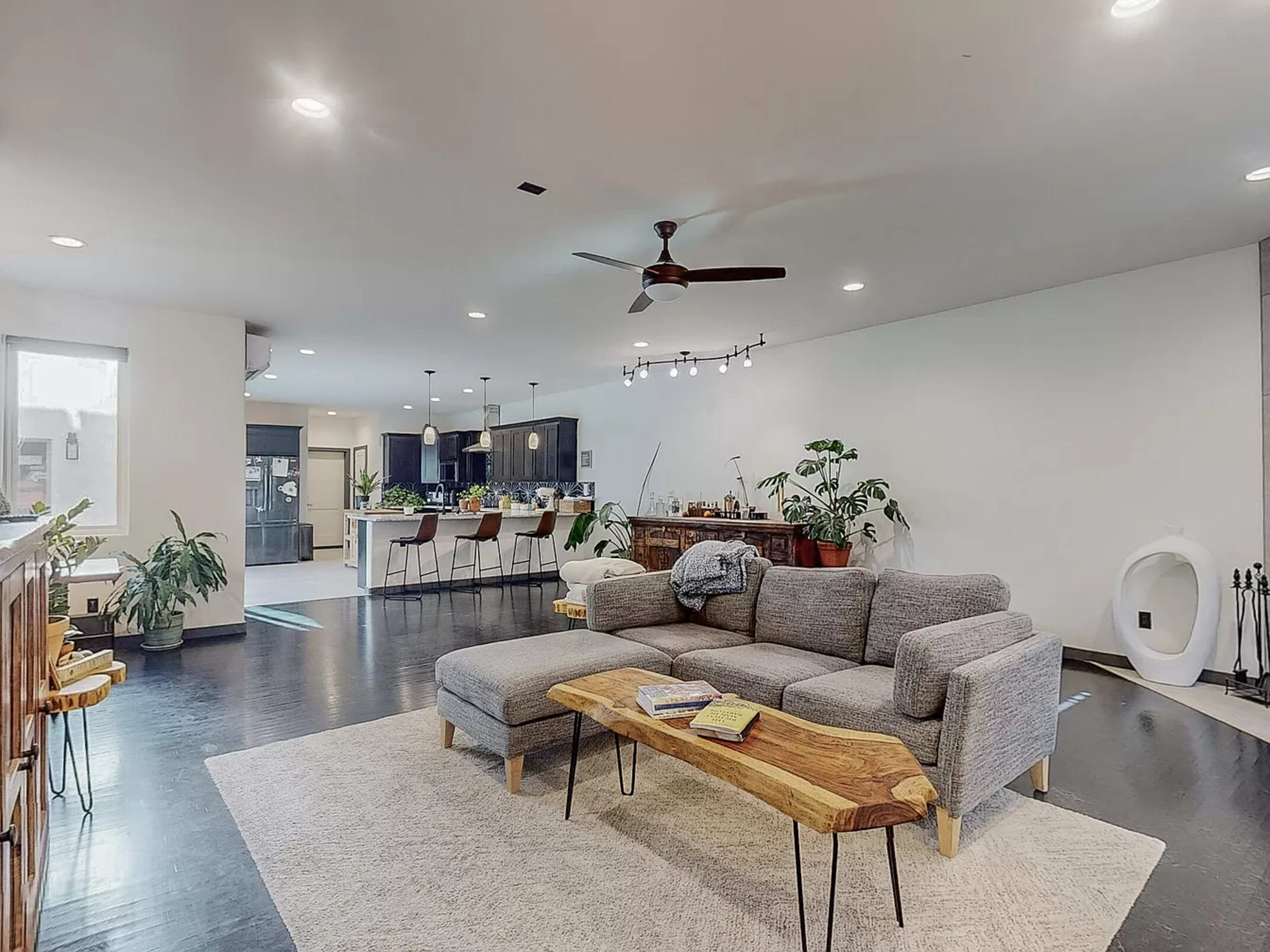 An open-concept living area featuring a grey sectional sofa, a natural wood coffee table, and a kitchen with bar stools in the background.