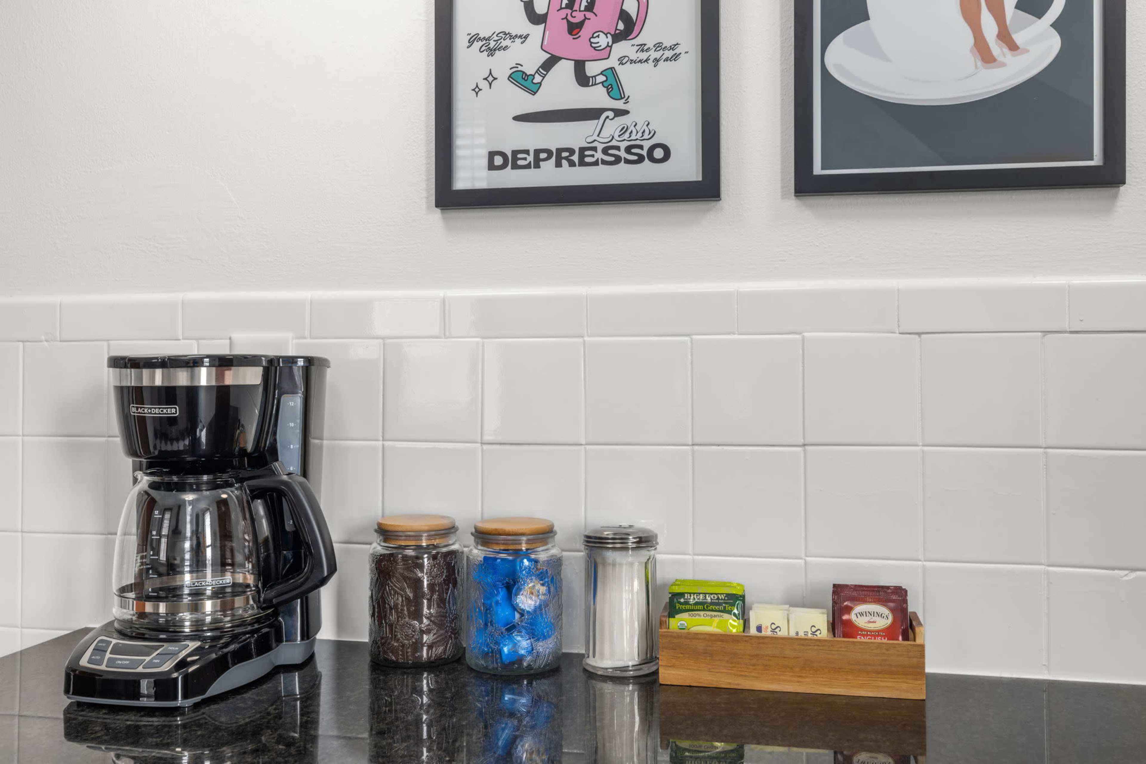 A coffee maker is placed on a countertop next to jars of sugar, coffee, and tea bags, with framed art on the wall above.