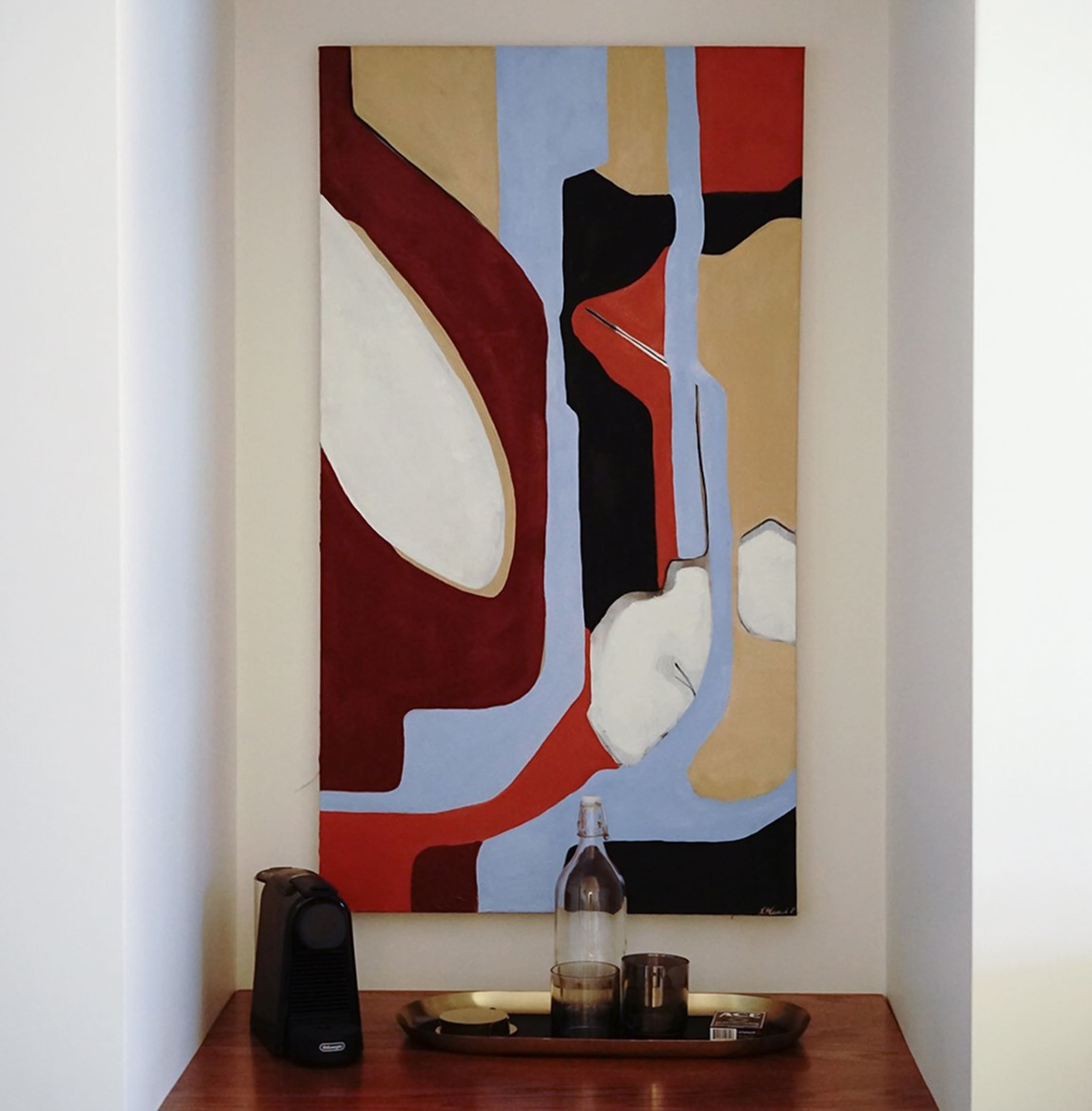 A colorful abstract painting hangs on a wall above a wooden surface with a water bottle, a glass, and a small appliance.