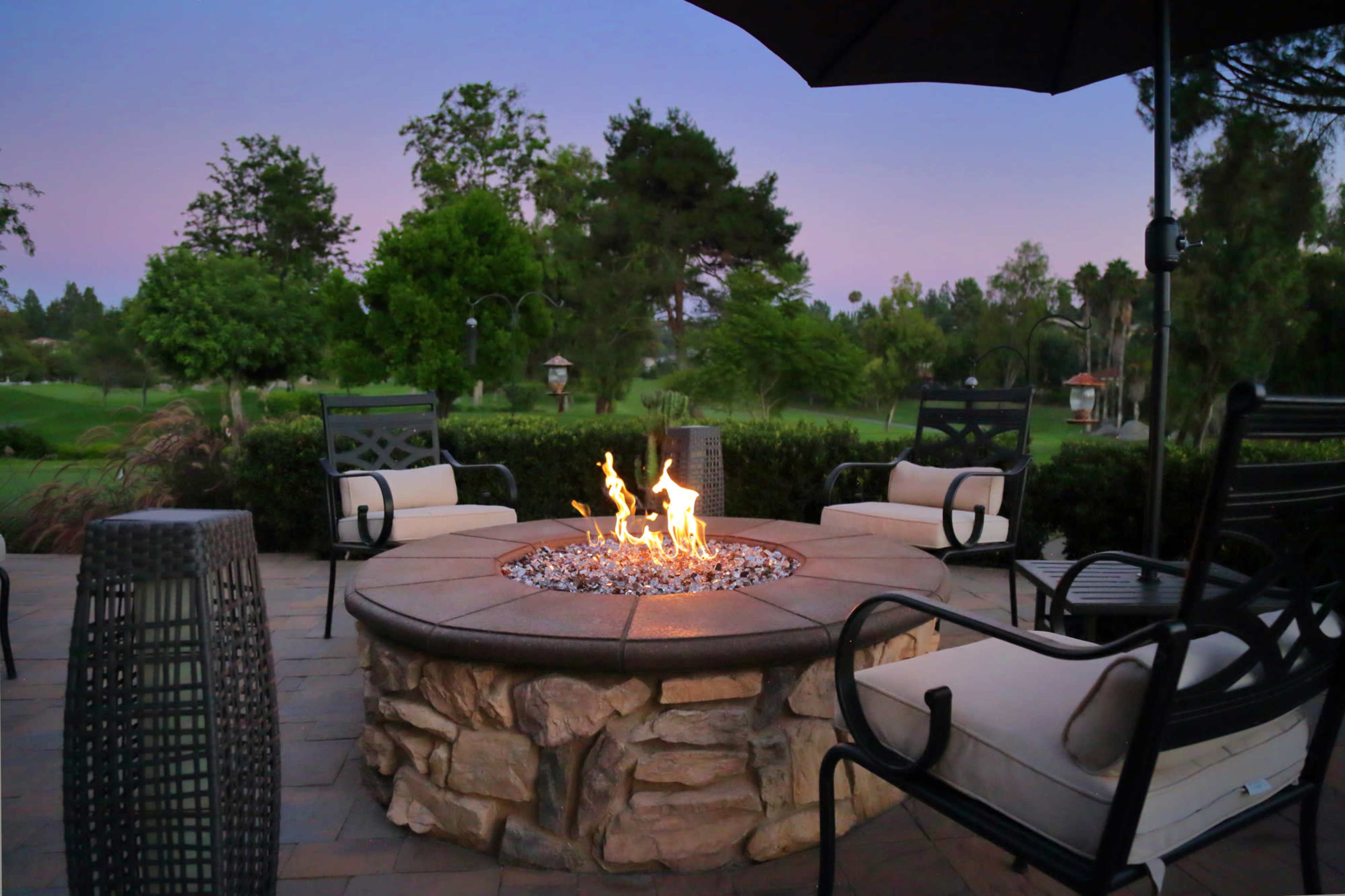 A stone fire pit with flames is surrounded by four chairs on a patio overlooking a green landscape at dusk.