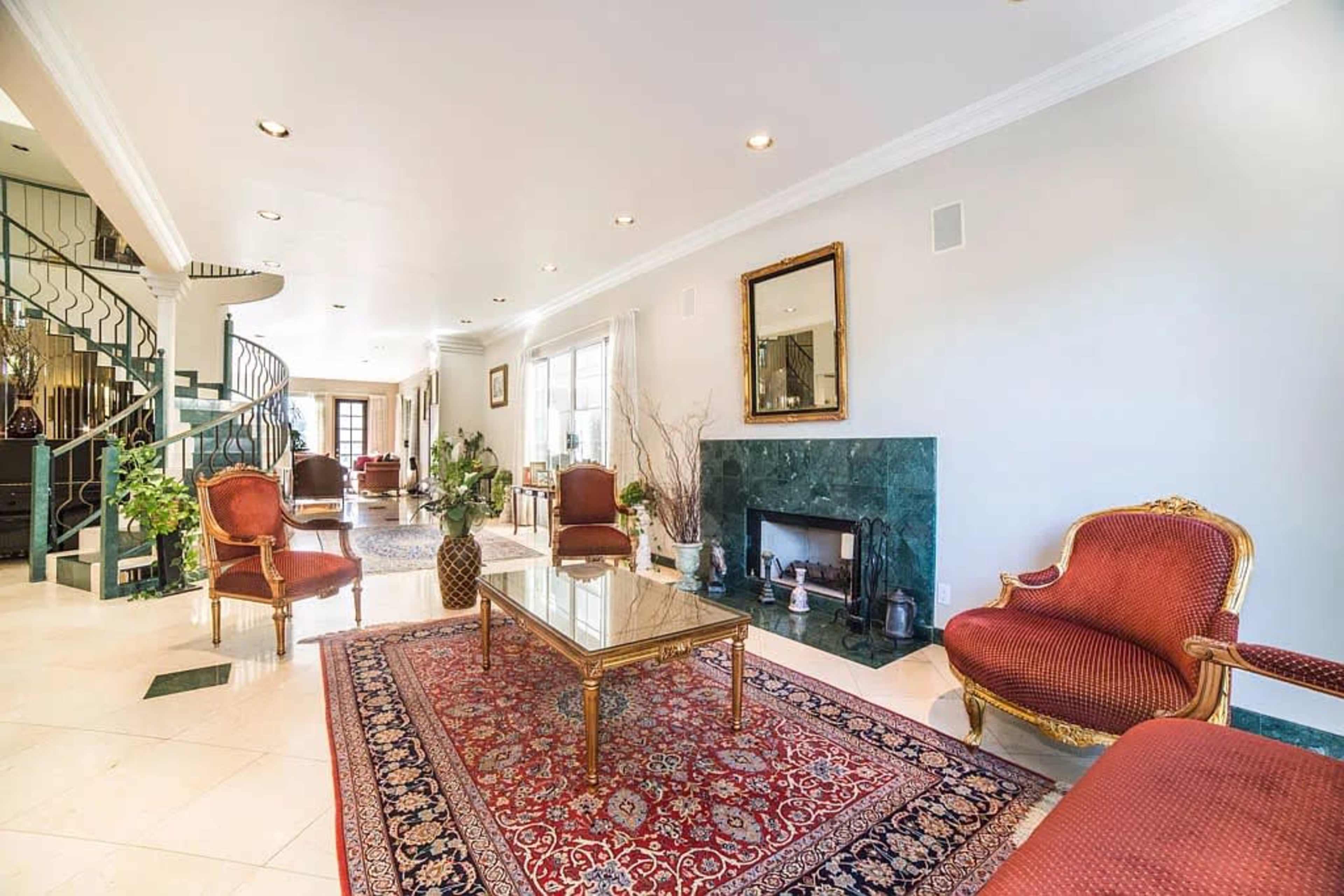 The image shows a spacious living room with cream-colored walls, a green marble fireplace, a patterned area rug, and red upholstered chairs arranged around a glass coffee table.