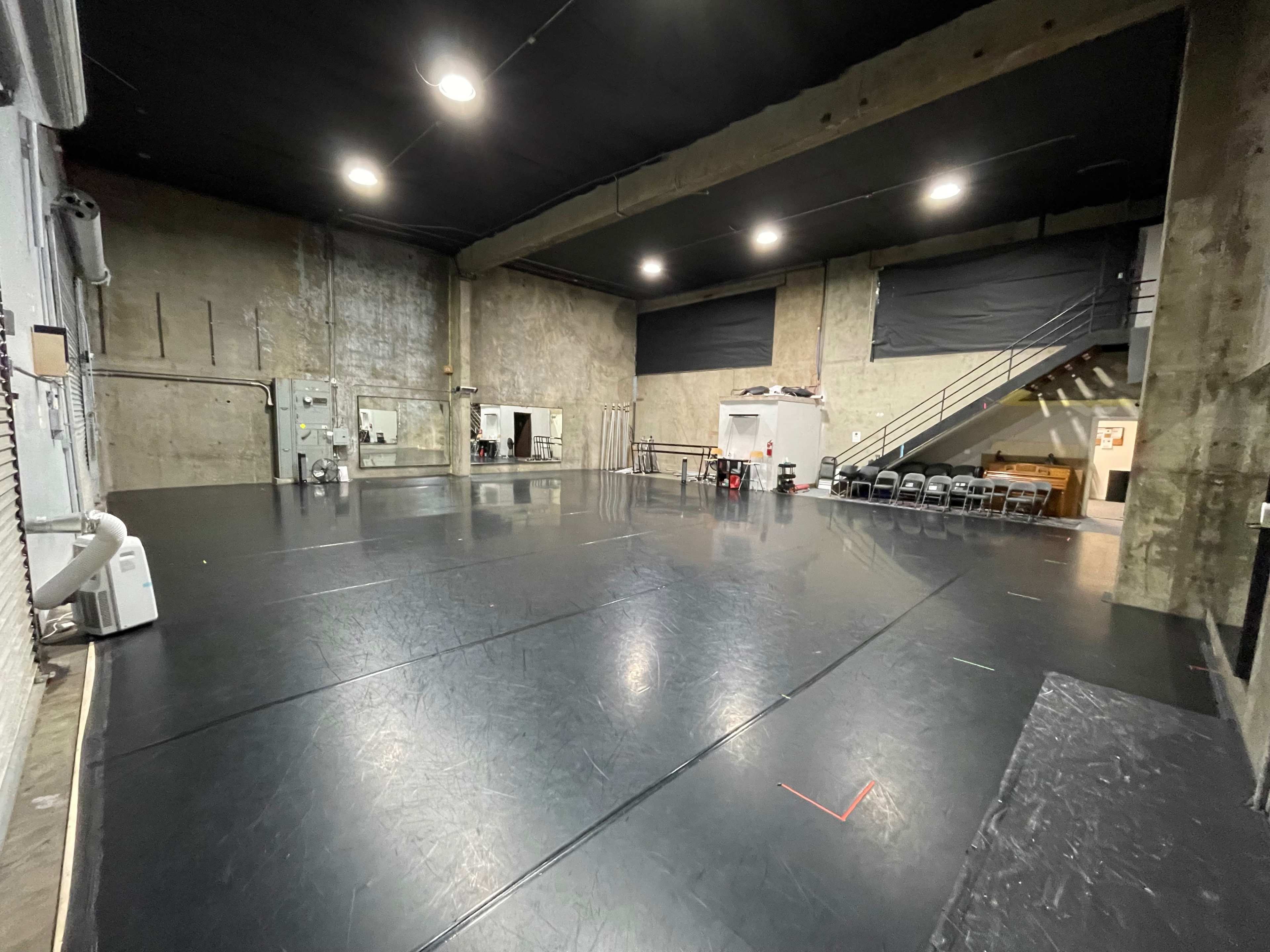 The image shows a spacious dance studio with a dark, polished floor, concrete walls, and a staircase in the background leading to a seating area.