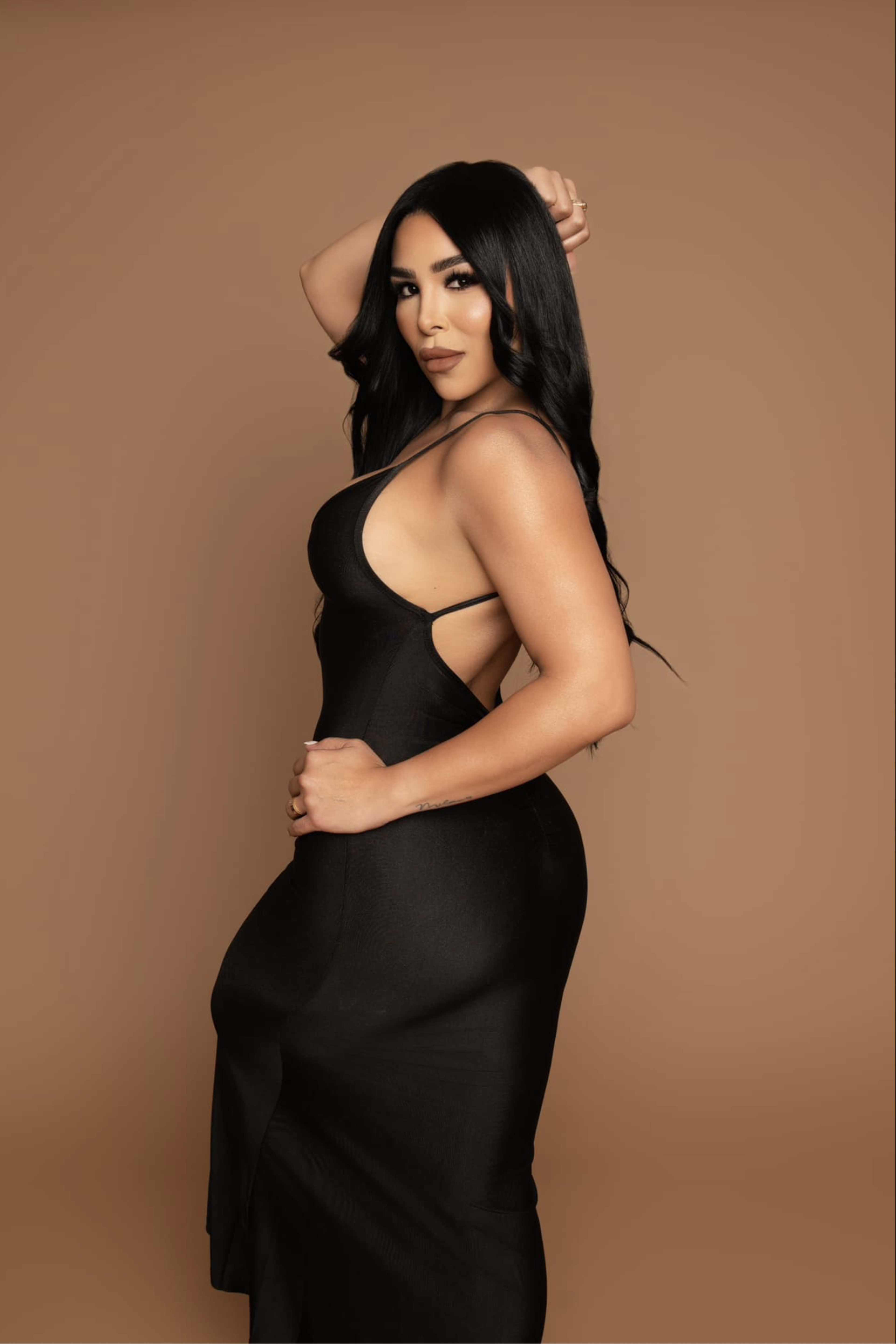 A woman in a form-fitting black dress poses against a warm brown background.