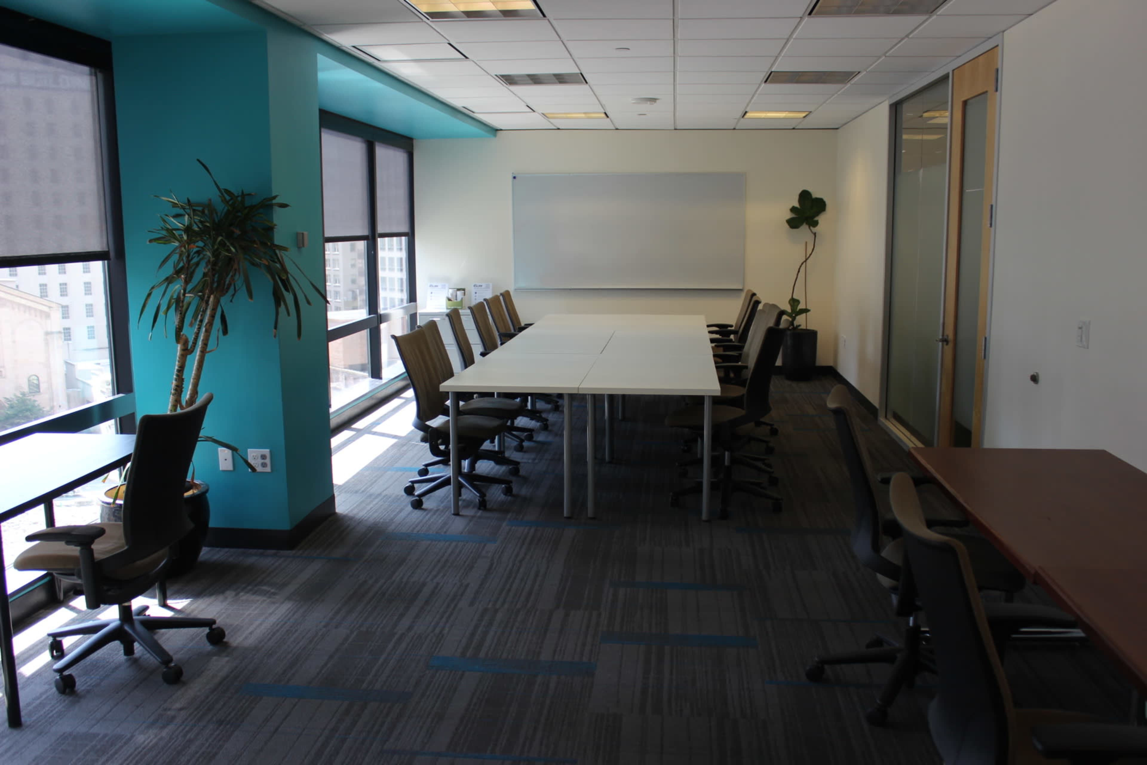 A conference room features a long table with several chairs, large windows, and potted plants along the walls.