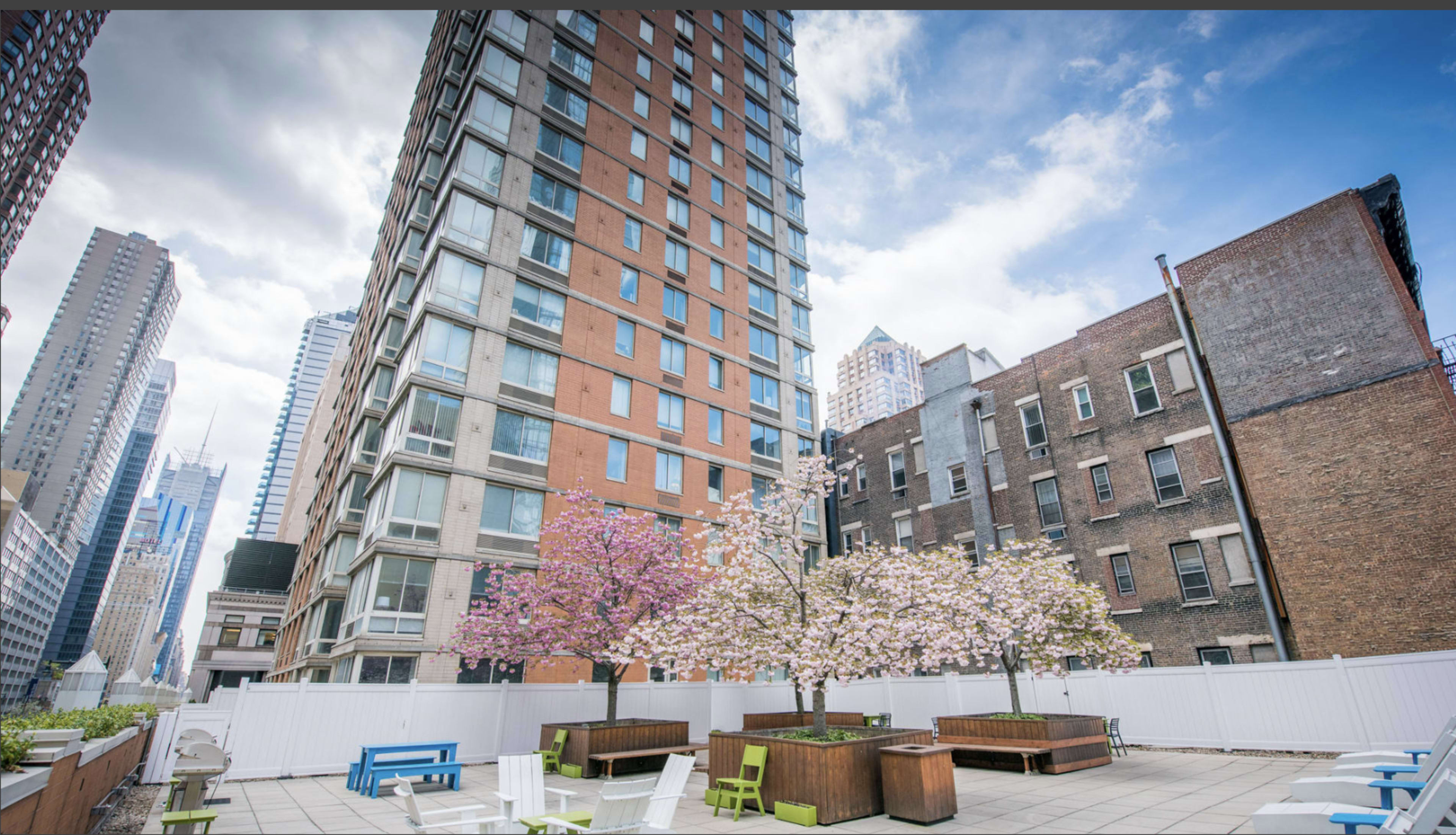 The image shows a rooftop area in an urban setting with modern buildings, featuring cherry blossom trees and outdoor seating.